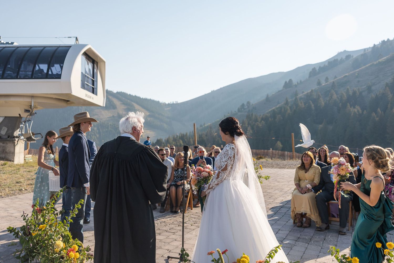 A mountain wedding ceremony takes place at a ski resort with guests seated outdoors on a sunny day.