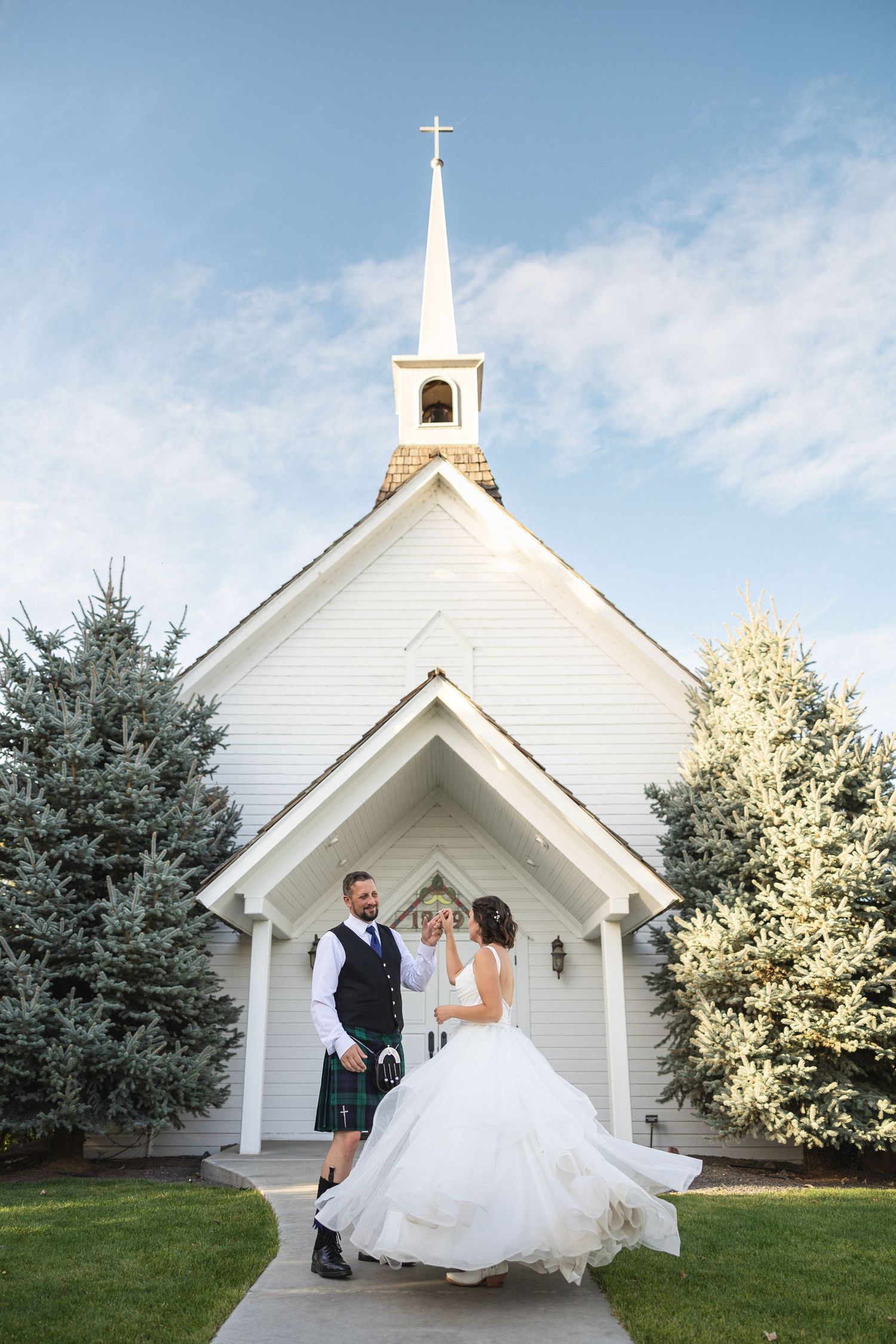 A couple dancing together in front of a white chapel with evergreen trees on either side.