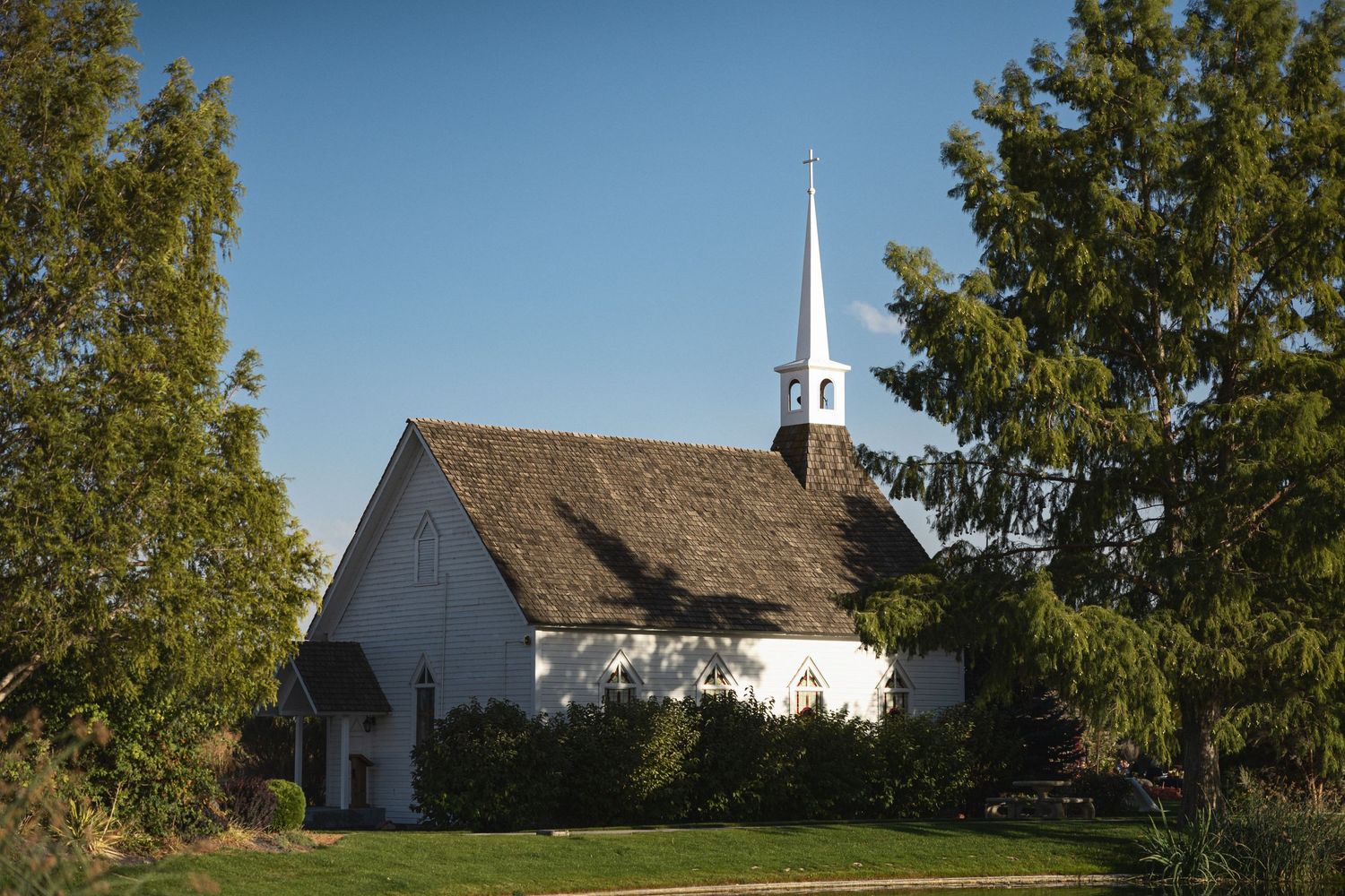A charming white country church with a tall steeple set against blue skies and framed by green trees.
