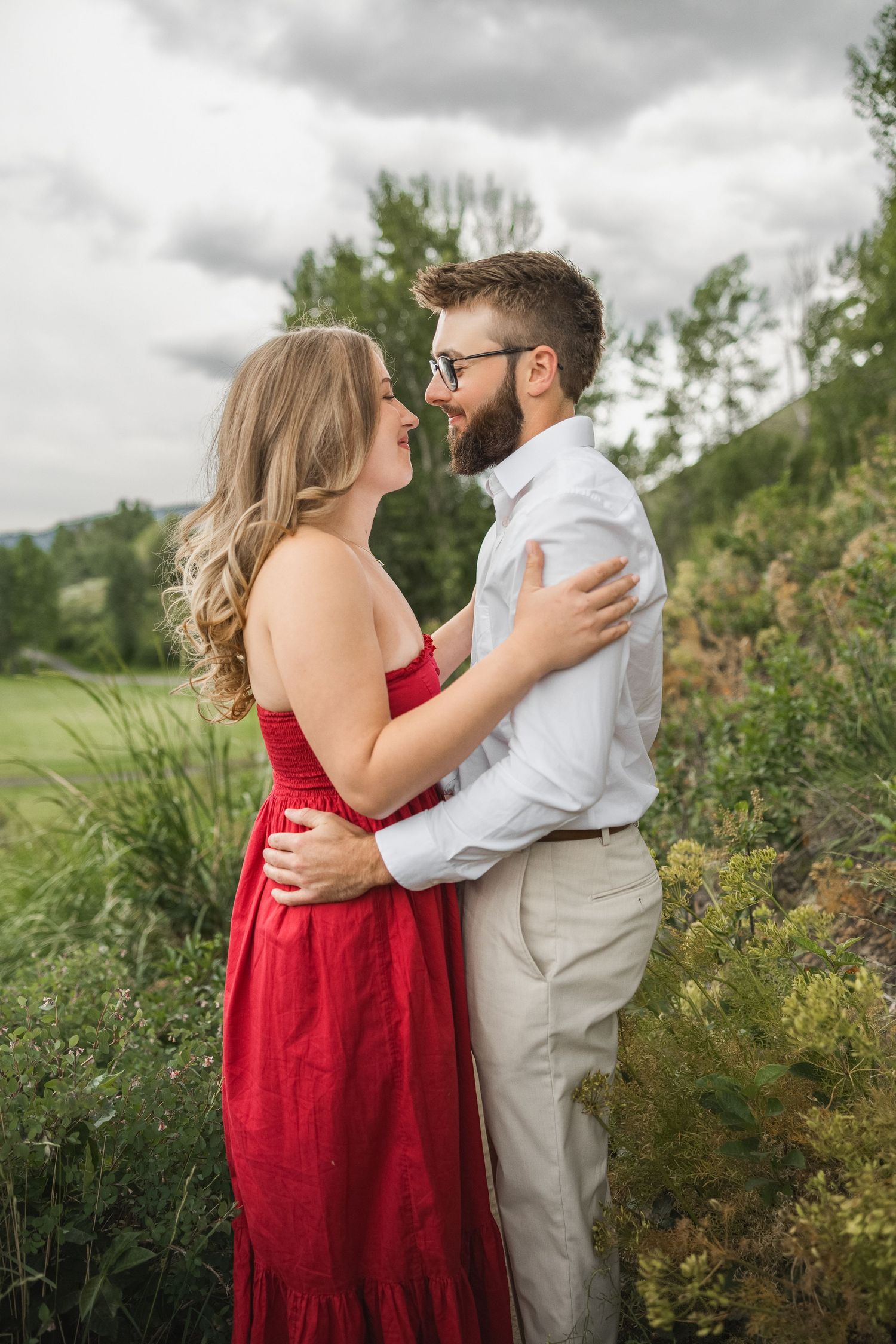 A romantic couple in a red dress and white shirt embrace in a lush outdoor setting with cloudy skies.