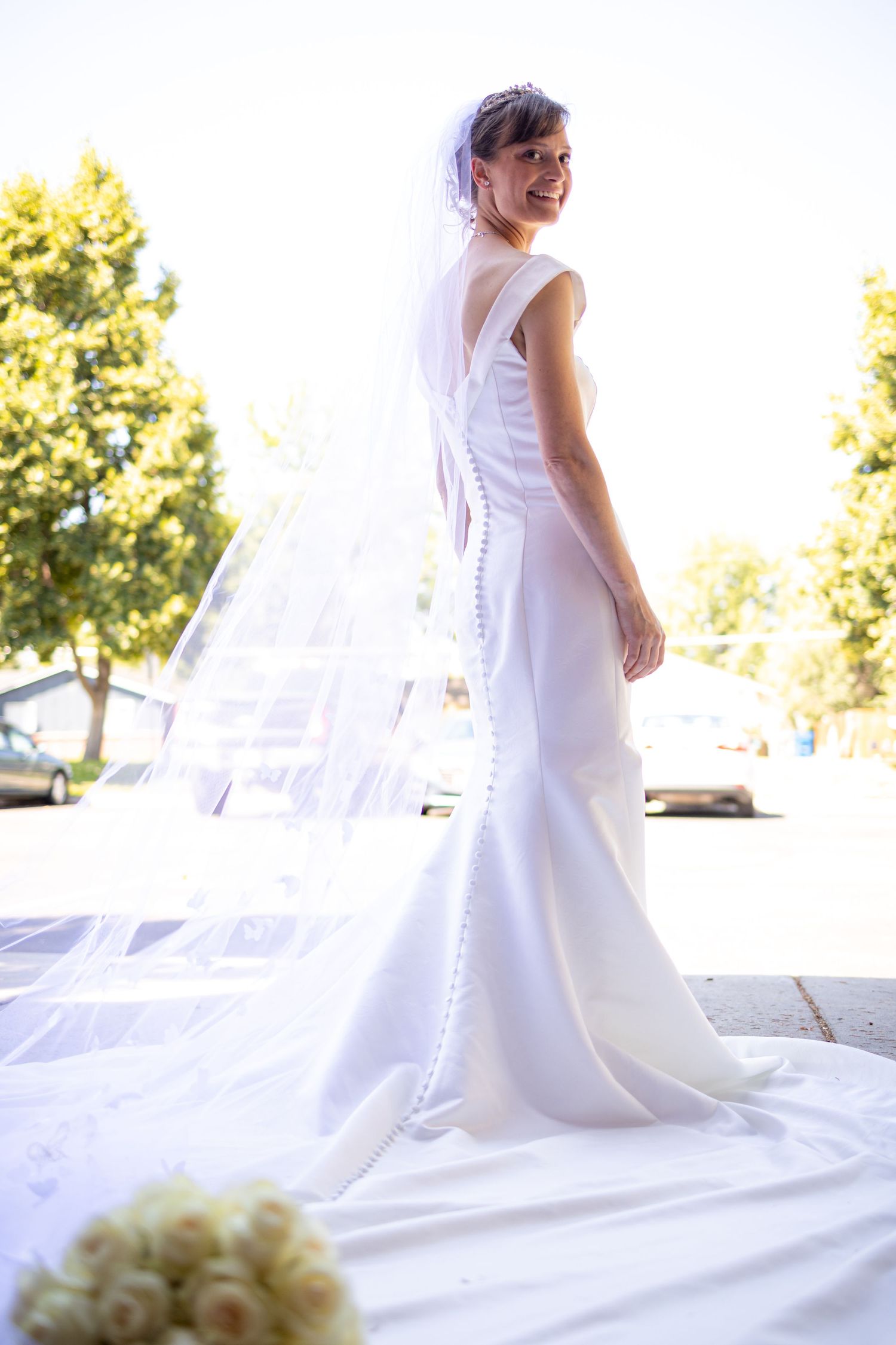 Elegant bride in fitted white dress and veil poses outdoors with bouquet on ground.