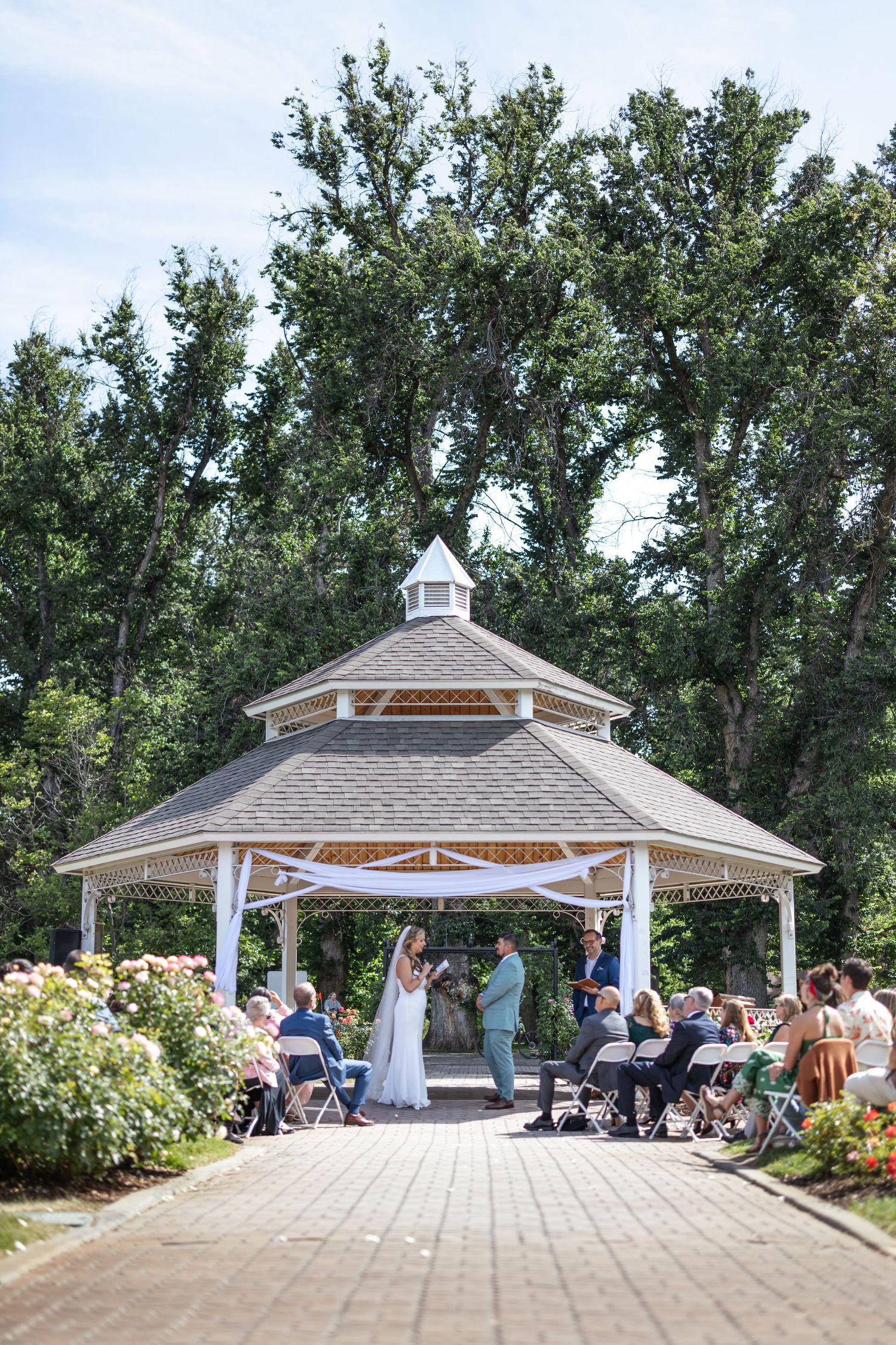 Wedding ceremony in white gazebo surrounded by trees and garden landscape.