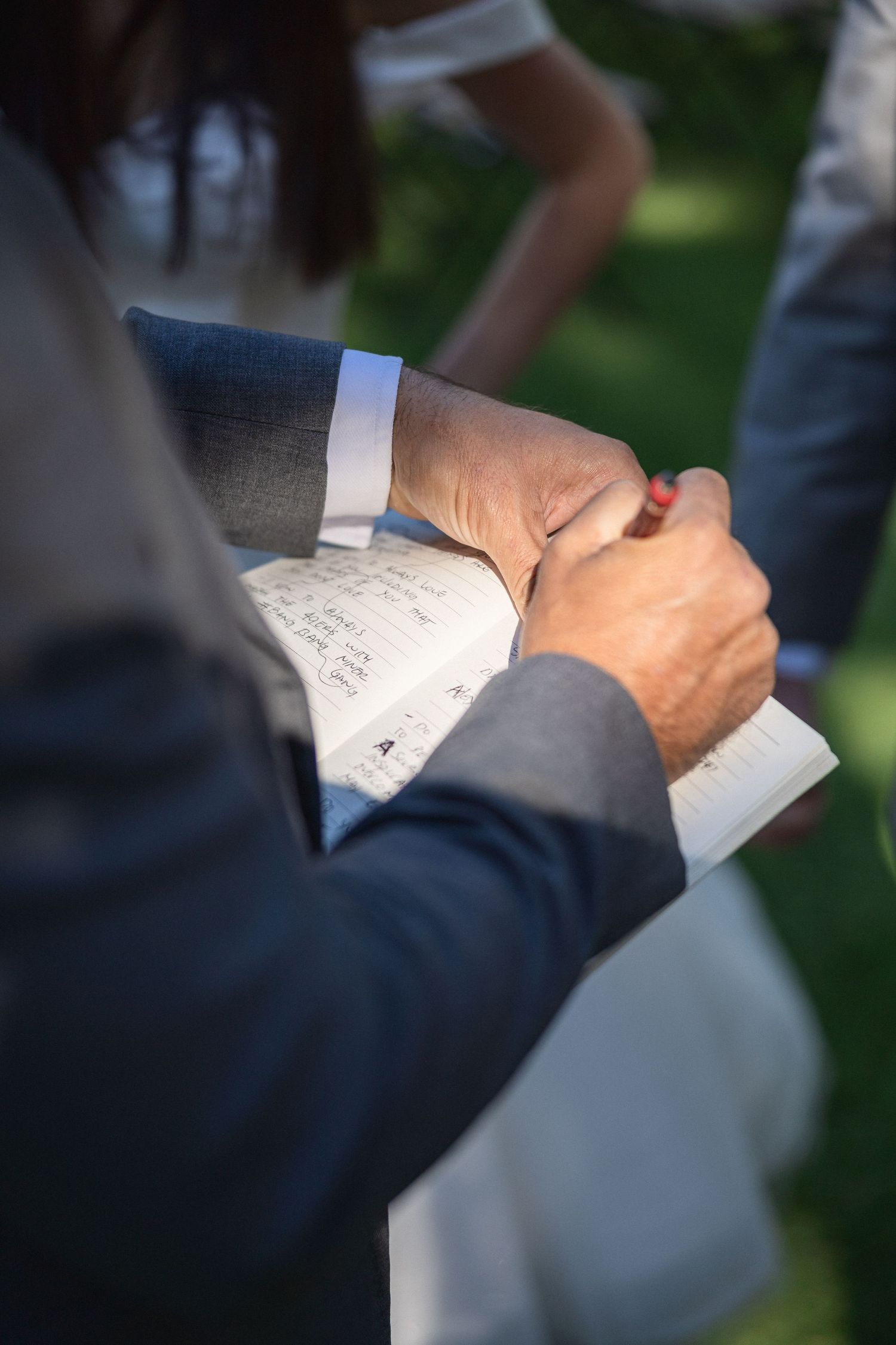 Close-up of hands writing in a notebook during outdoor wedding ceremony.