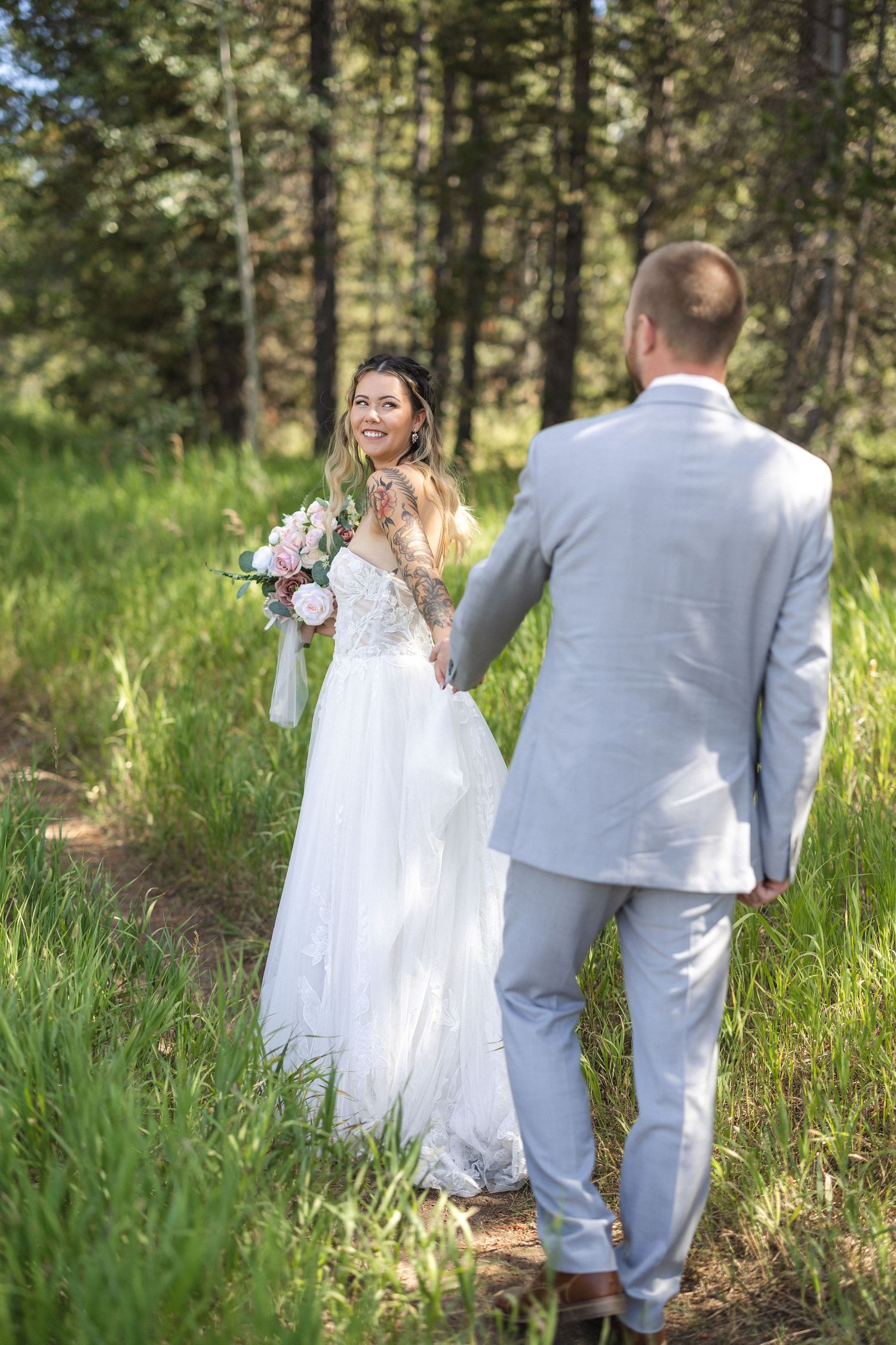 Wedding couple walks hand in hand through tall grass in a forest setting.