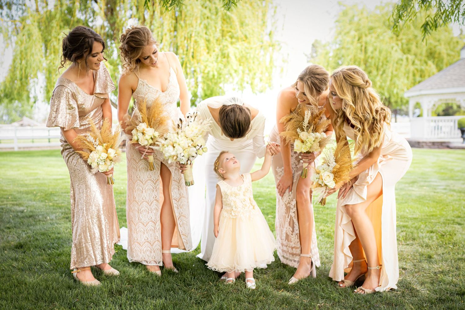 Bridesmaids in blush dresses interact with flower girl on grassy lawn under willow tree.