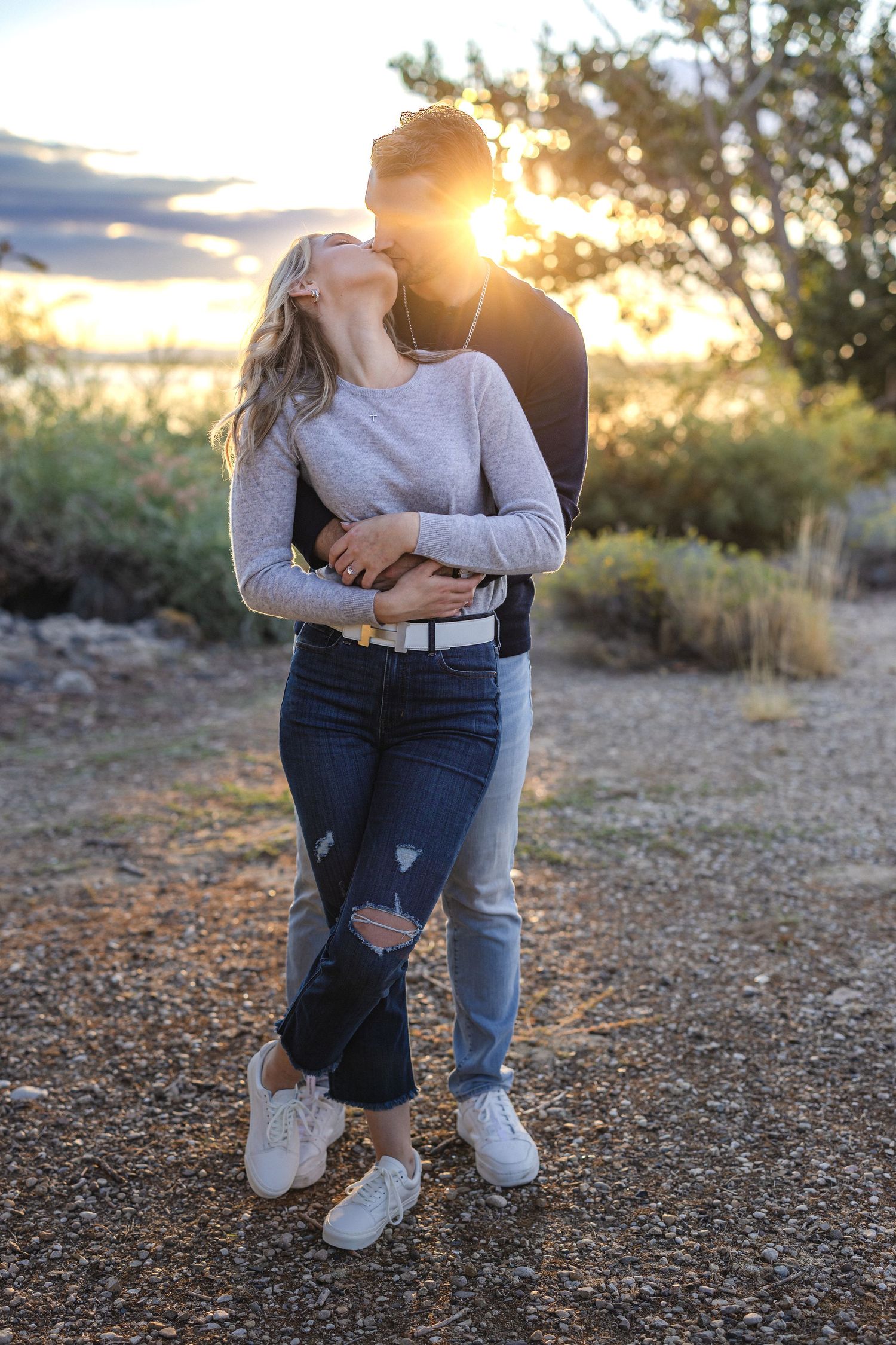 Couple in casual wear embracing outdoors during sunset with natural landscape backdrop.