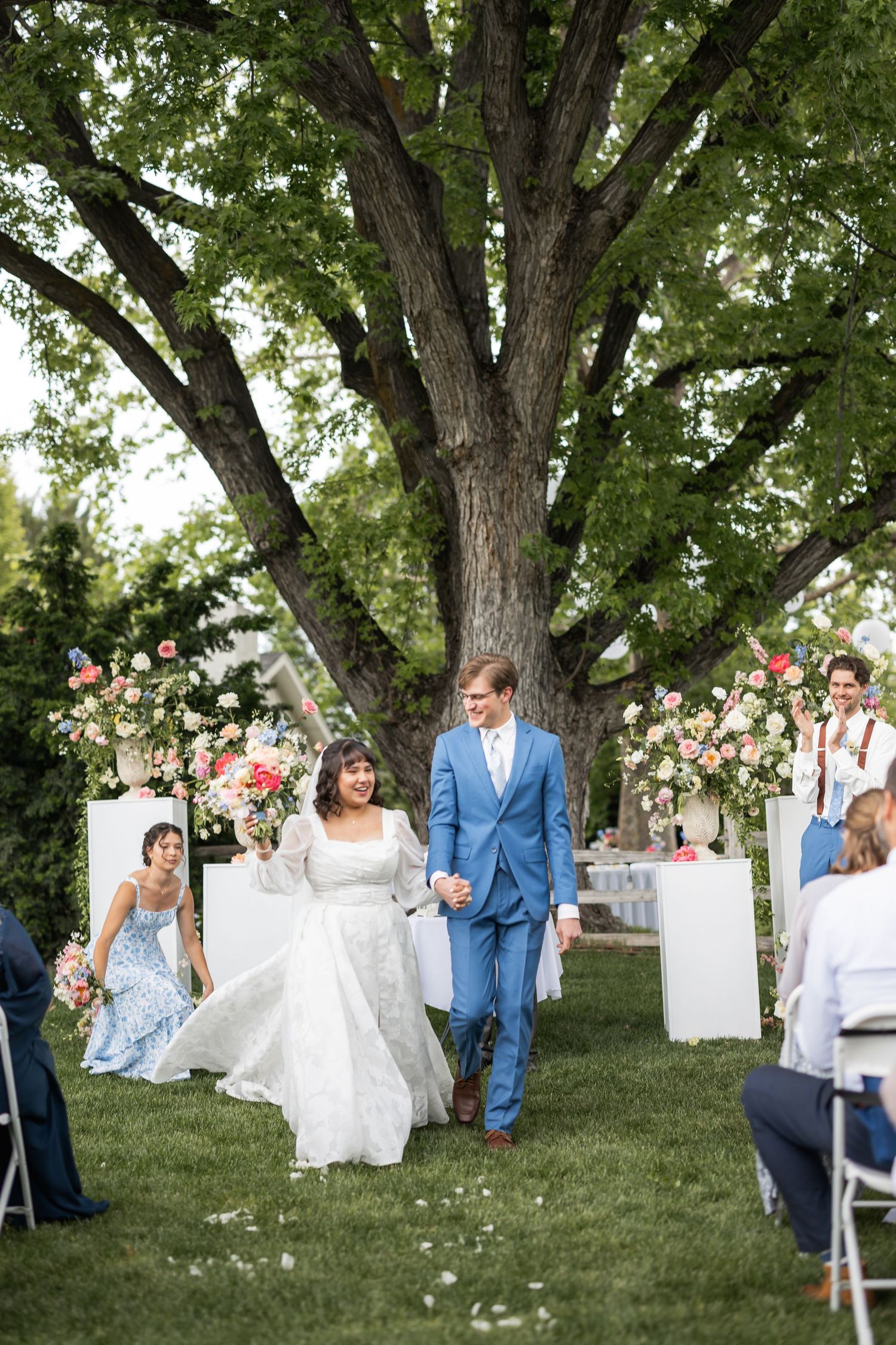 Couple walking down aisle under majestic tree with pink floral arrangements at outdoor wedding.