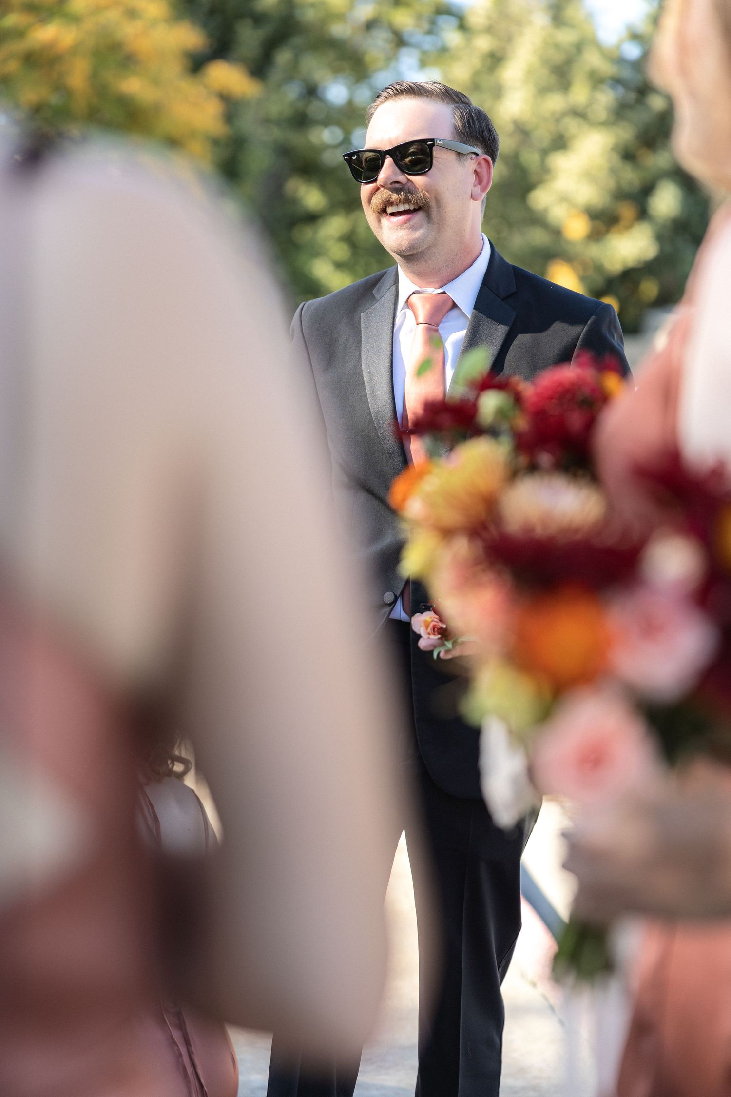 Wedding ceremony guest wearing sunglasses and suit with autumn florals in foreground.