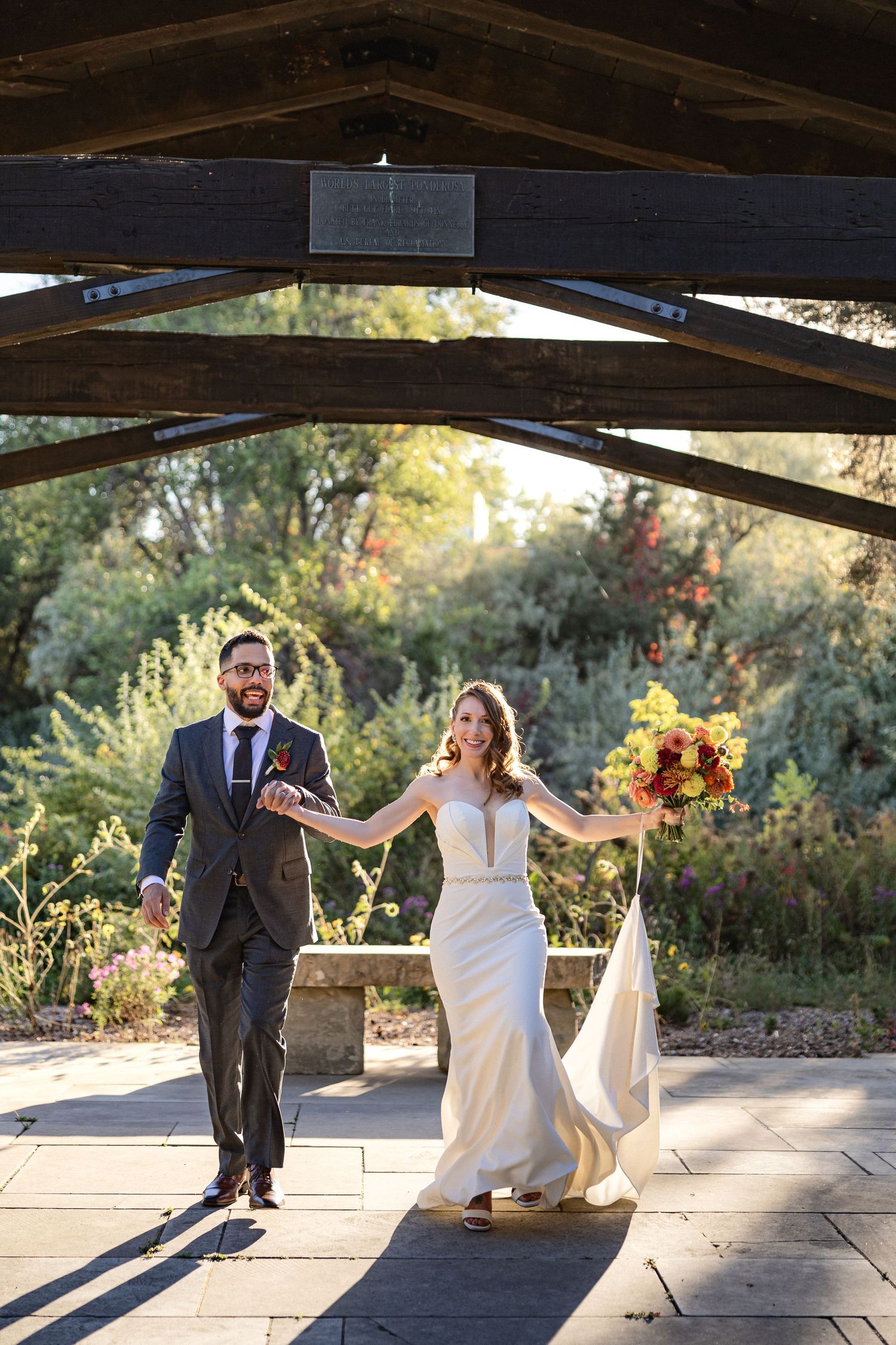 Happy newlyweds walk hand in hand under wooden pavilion in garden setting.