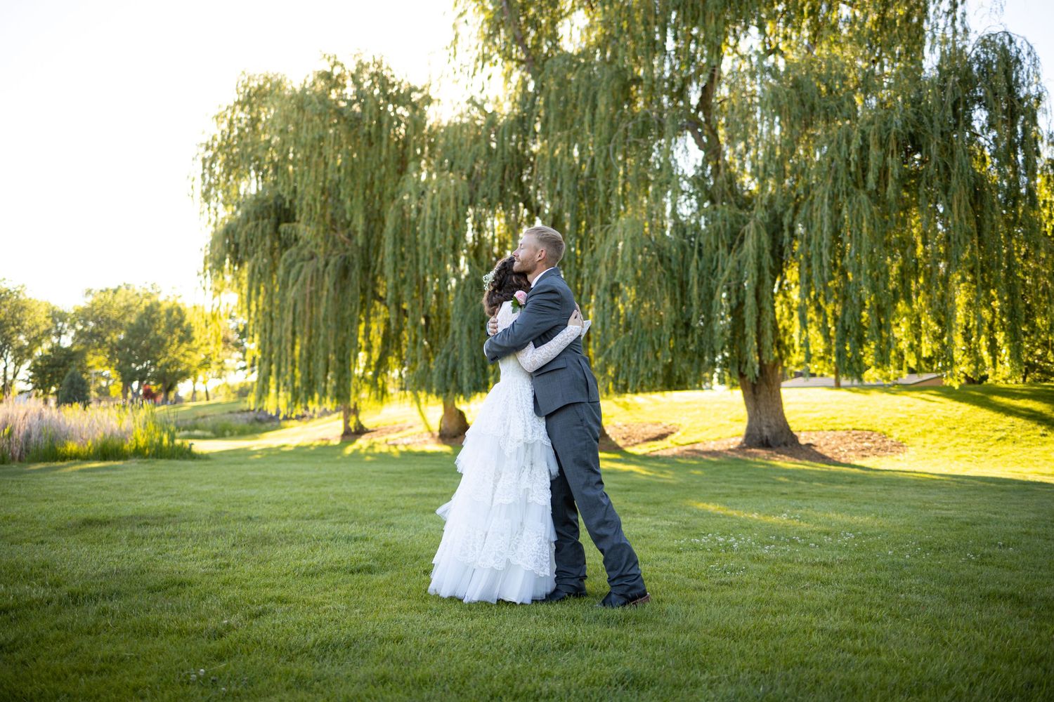 Wedding couple embraces on lawn with weeping willow trees creating romantic backdrop.