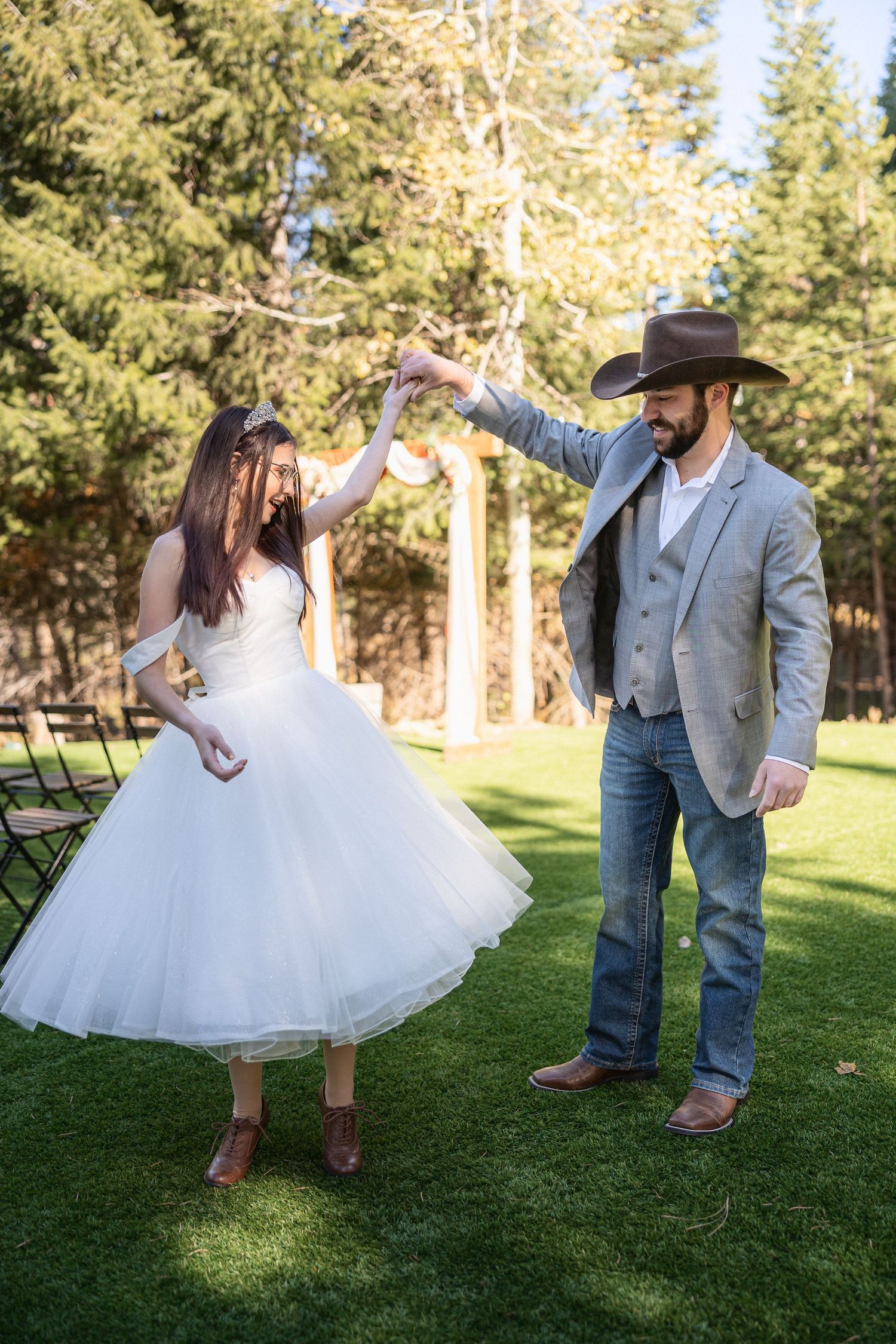 A bride and groom perform a joyful dance routine on green grass with trees in the background.