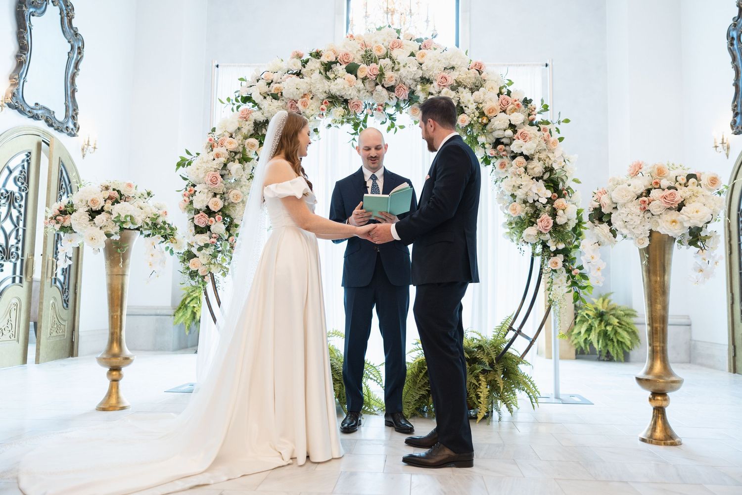 Elegant white wedding ceremony space decorated with floral arch and crystal chandeliers.