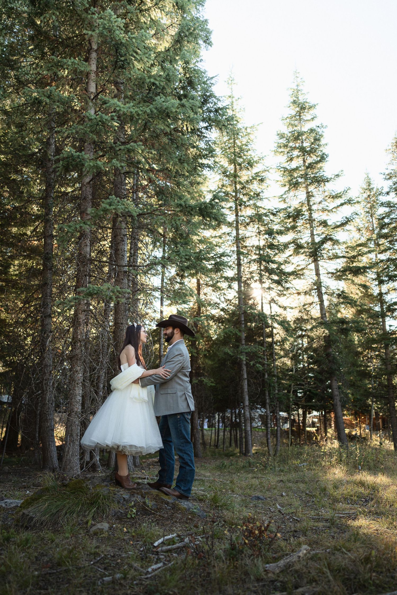 Bride and groom holding hands in a sunlit pine forest, groom wearing cowboy hat and jeans, bride in white tulle dress.