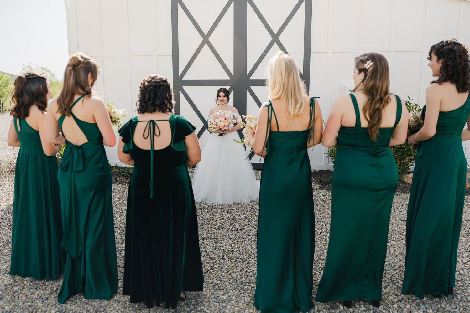 Six bridesmaids in emerald green gowns stand with their backs to the camera facing a bride in a white wedding dress.