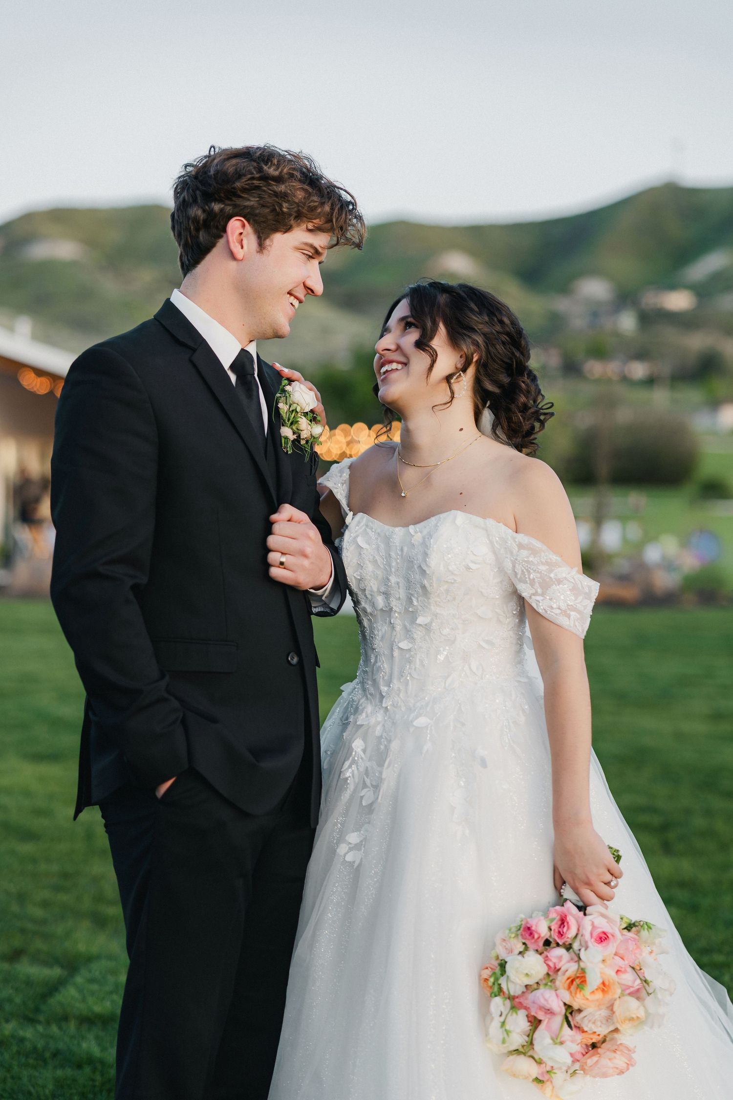 Bride and groom smiling at each other outdoors, she holds pink bouquet in white gown, he wears black suit.