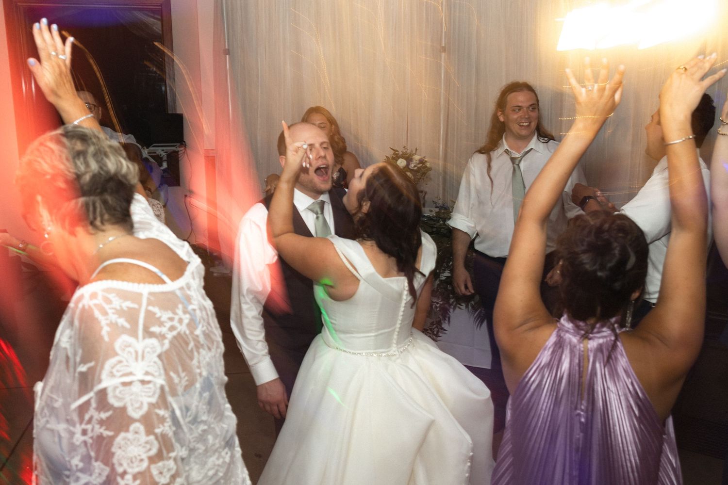 Wedding guests dancing and celebrating energetically on a colorfully lit dance floor.