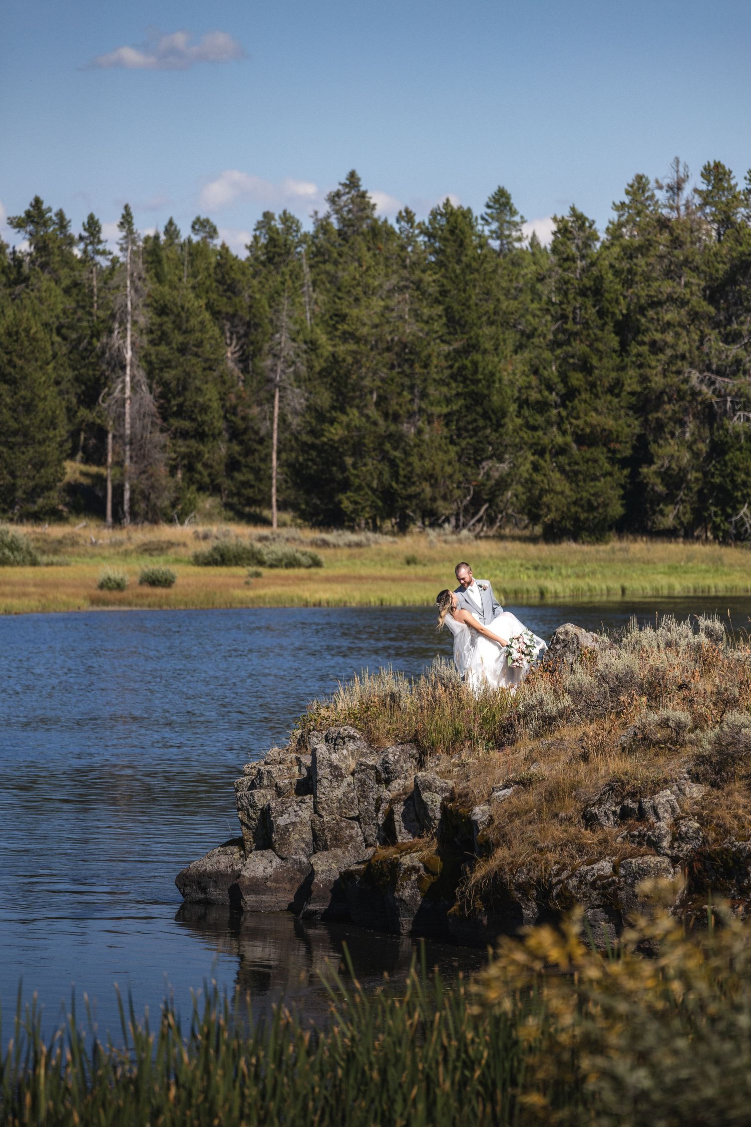 Wedding couple photographed atop rocky cliffside with evergreen forest and lake view.