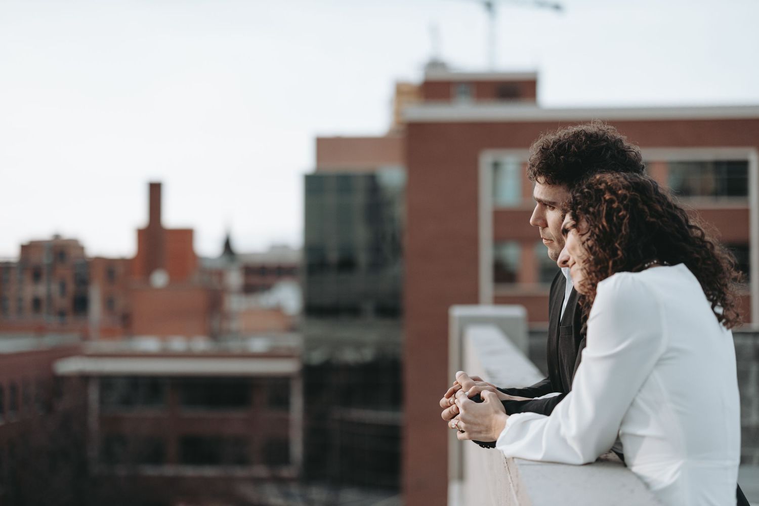 Person in white blazer looks thoughtfully over city rooftops while holding a drink.
