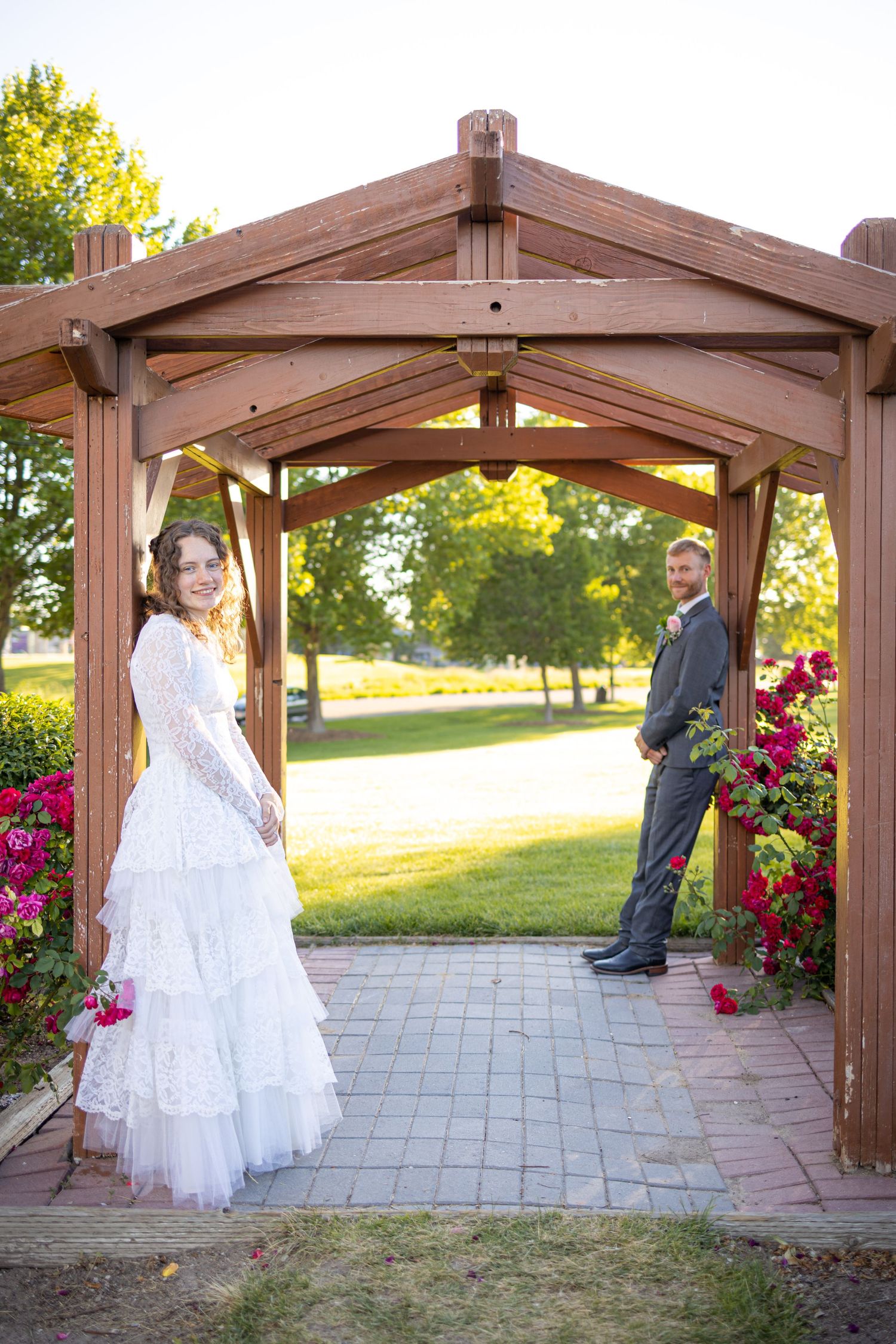 Wedding couple poses beneath rustic wooden archway adorned with red climbing flowers.