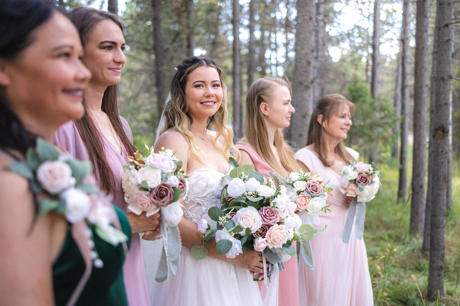 Bridal party smiling while holding pink and white bouquets in sunlit forest setting.