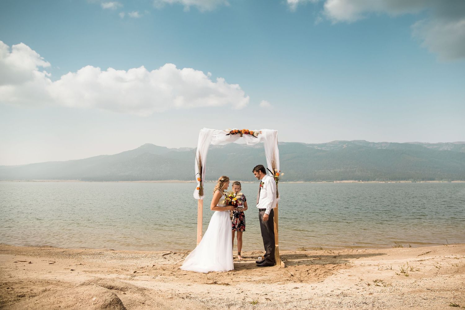 Wedding ceremony under an arch on a beach with mountains in the background.
