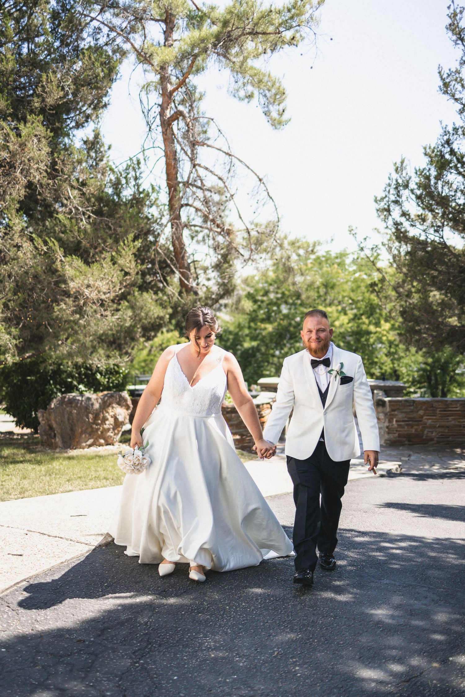A wedding couple walks hand in hand on a paved path surrounded by tall pine trees on a sunny day.