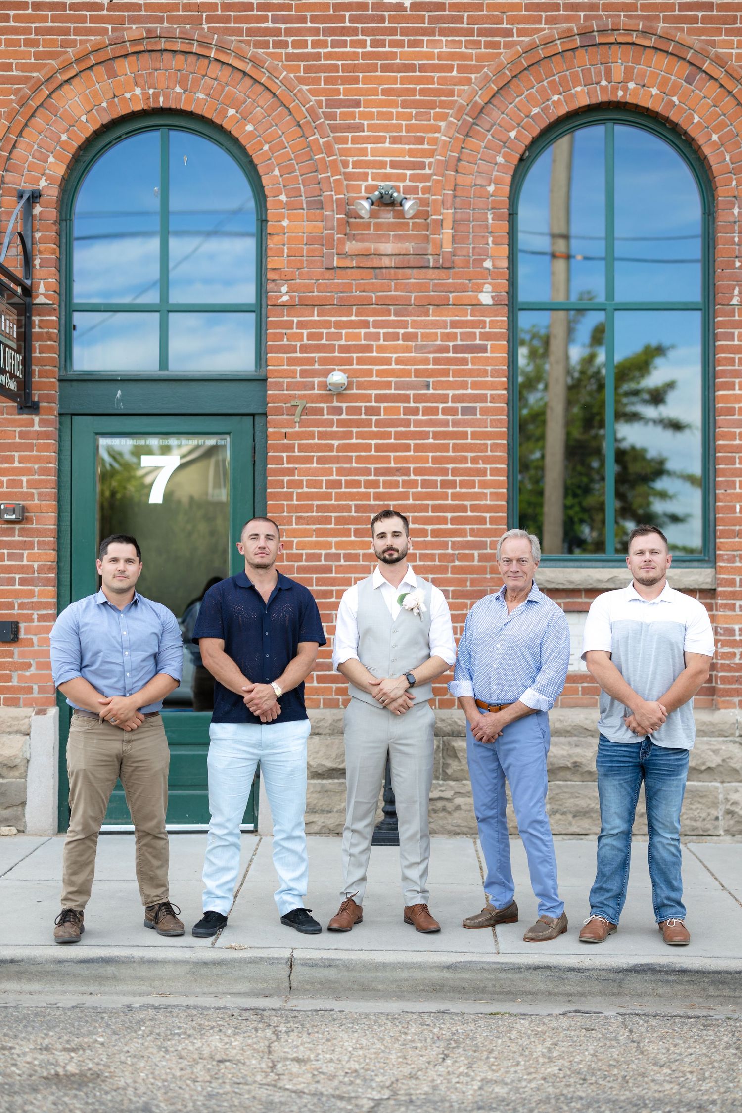 Groomsmen in casual attire stand in line on steps of brick building.