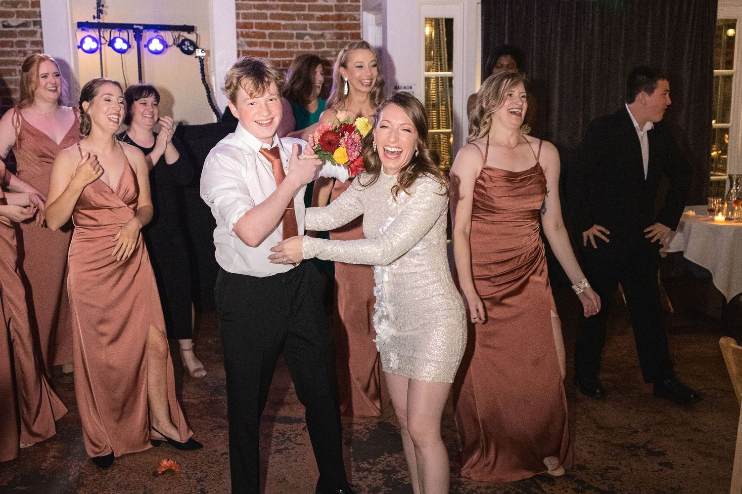 Wedding guests and bridal party dancing at a festive indoor reception with exposed brick walls.
