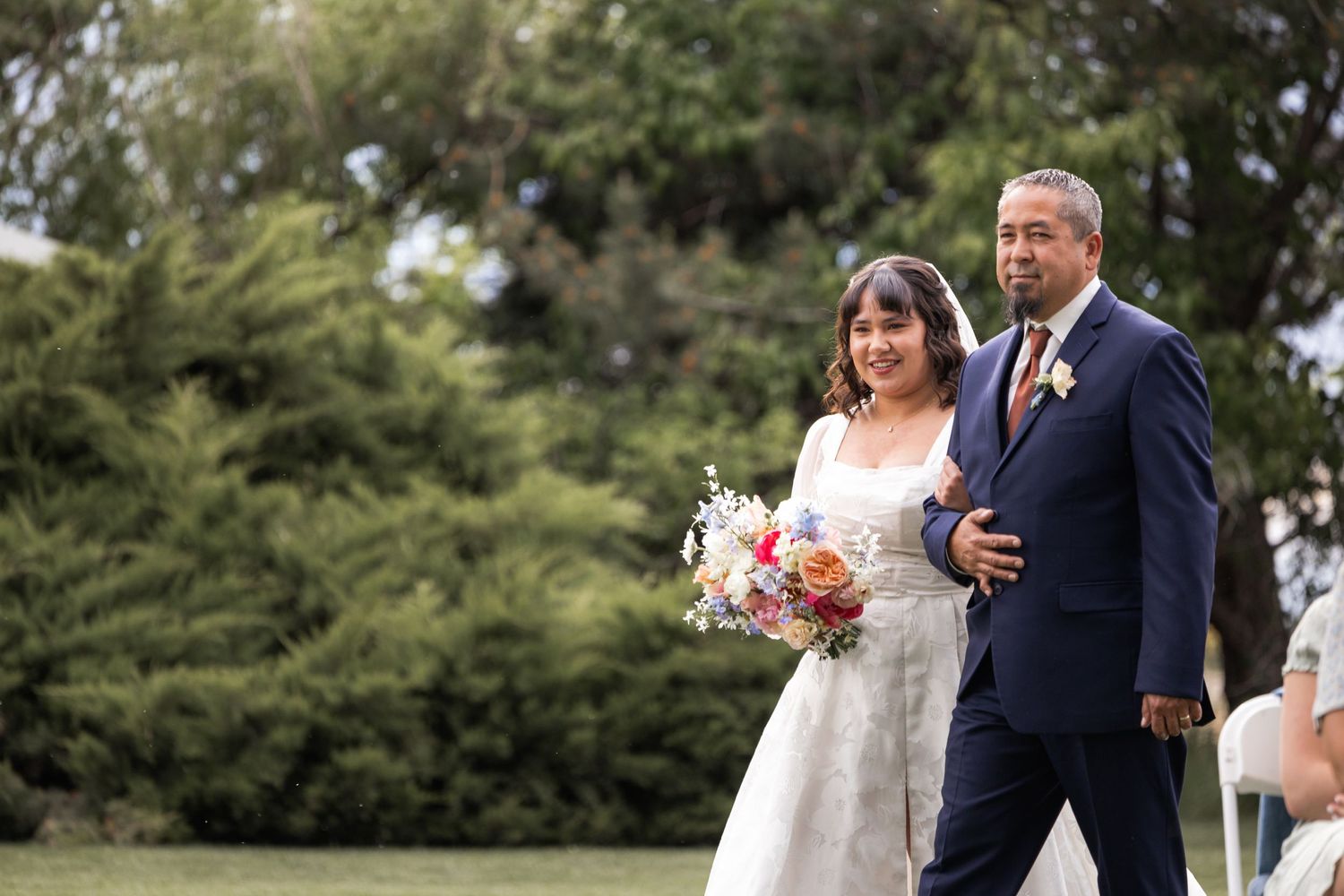 A couple walks down the aisle during an outdoor wedding ceremony with greenery in background.