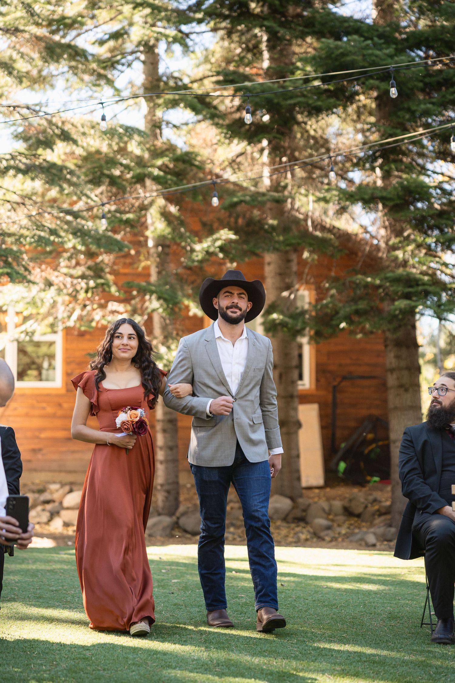 Wedding ceremony procession on a lawn with pine trees and rustic cabin backdrop.