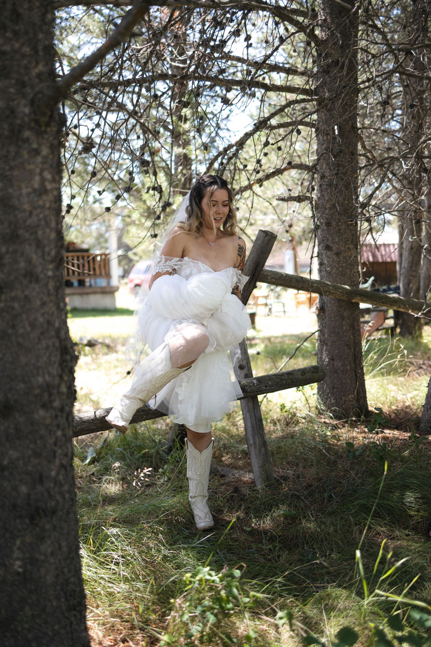 Bride in white dress and boots posing playfully on wooden fence among pine trees.