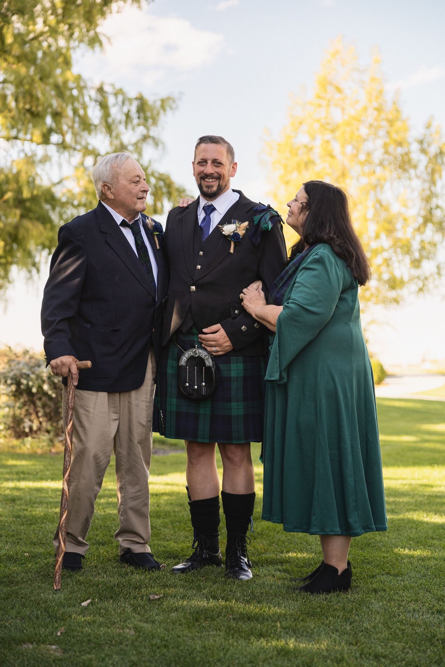 Wedding party members share a laugh outdoors with someone in traditional Scottish kilt attire.