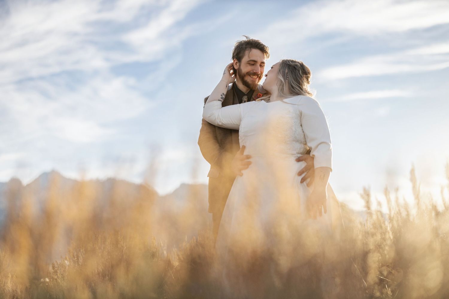 A couple embraces in a golden wheat field during sunset with mountains in the background.
