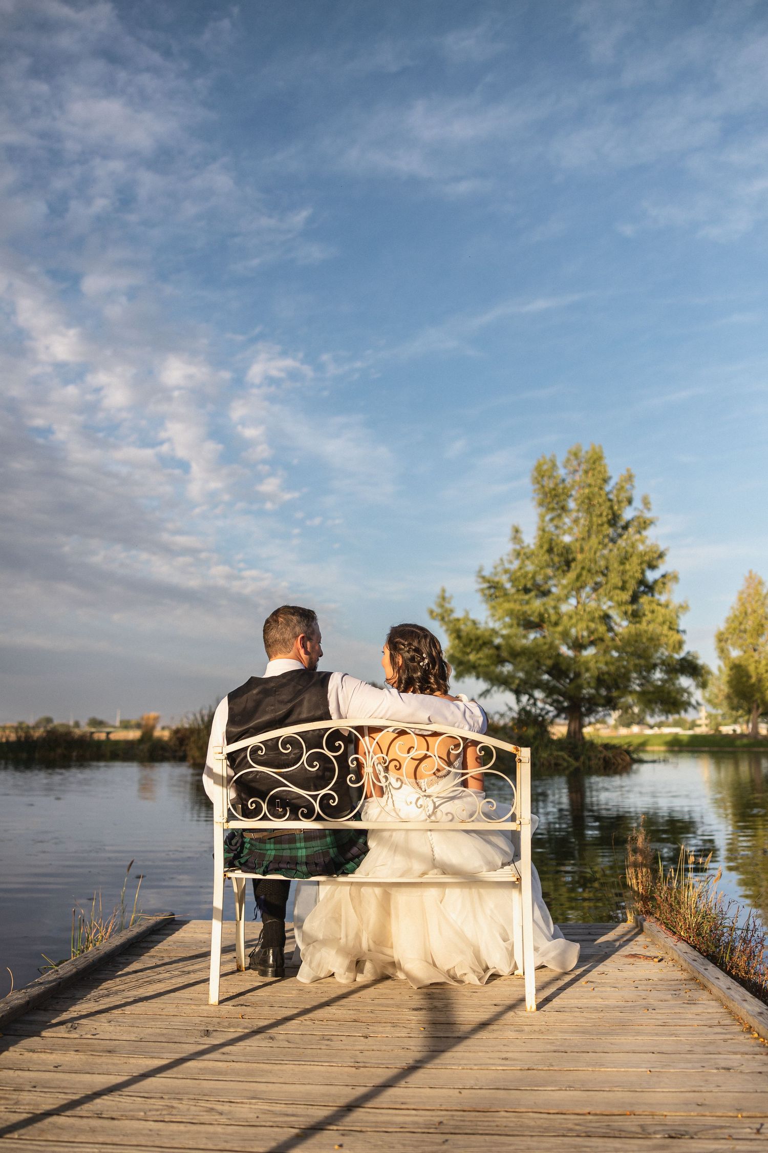 A couple sits together on a white bench overlooking a serene lake at sunset.