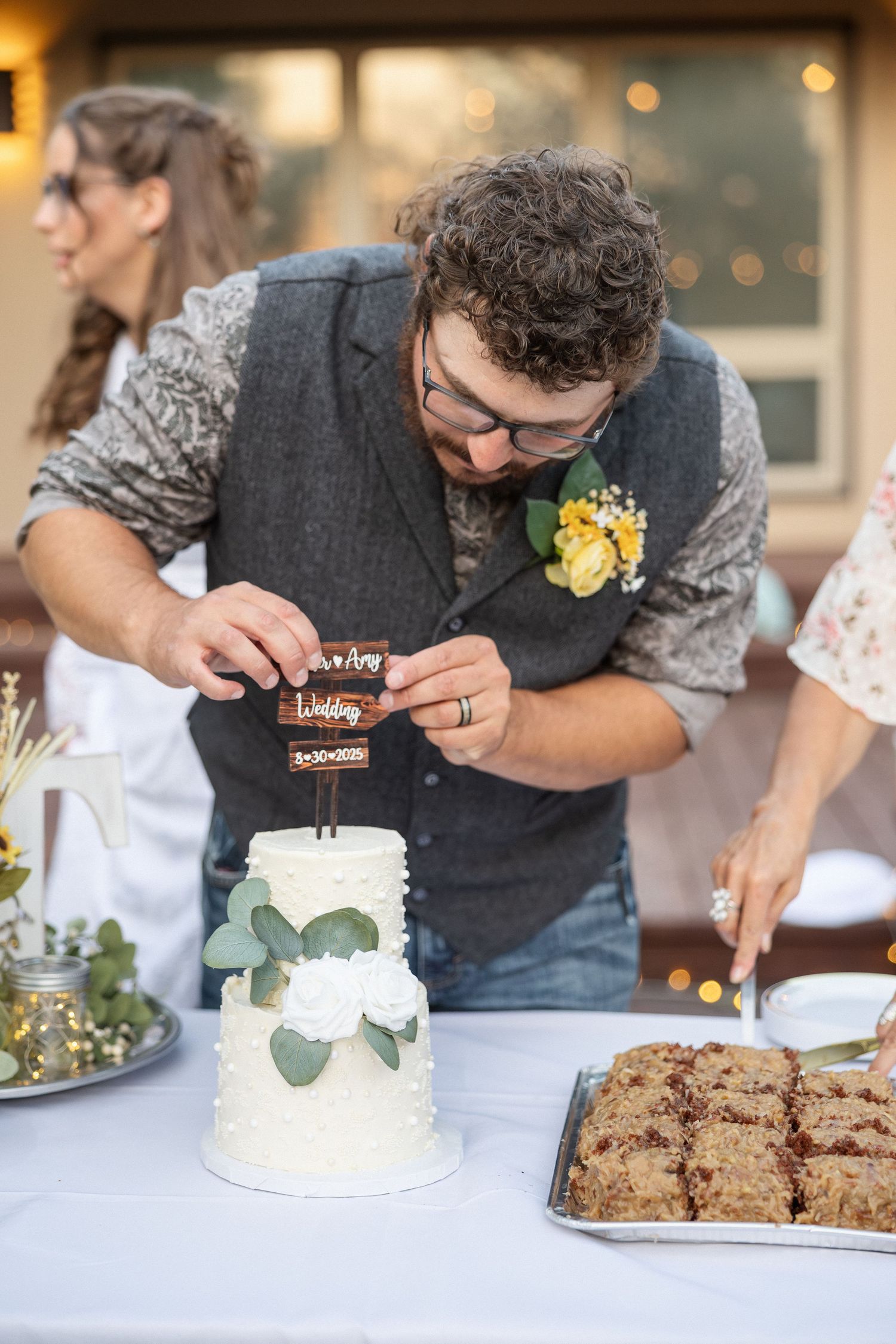 Couple cutting into a white wedding cake with yellow flowers and eucalyptus on a dessert table.