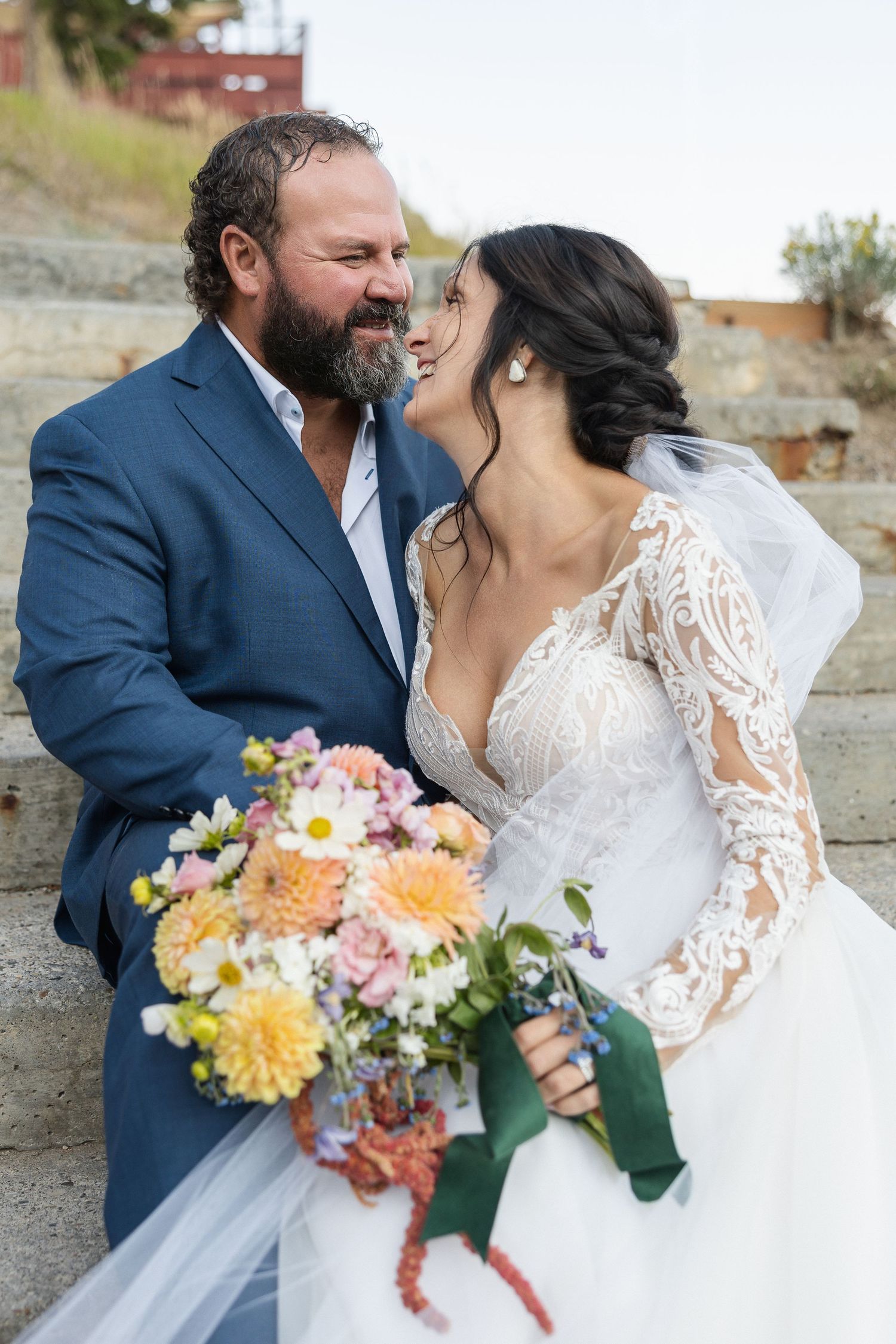A couple in wedding attire share intimate moments while holding a colorful bouquet of flowers against a rustic backdrop.