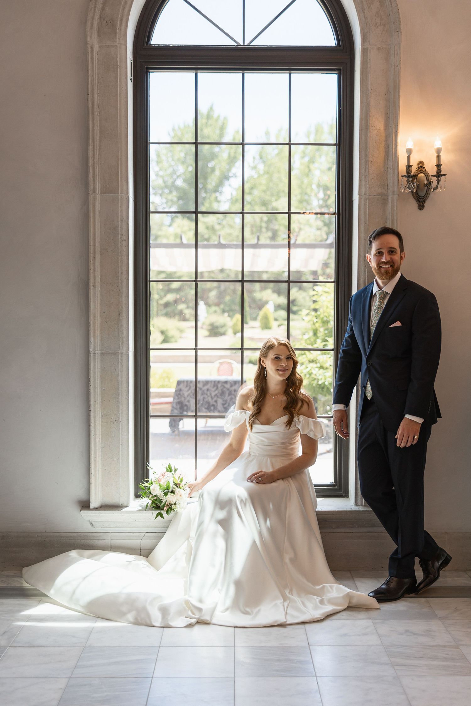 Wedding couple poses by large arched windows in an elegant indoor setting with natural light.