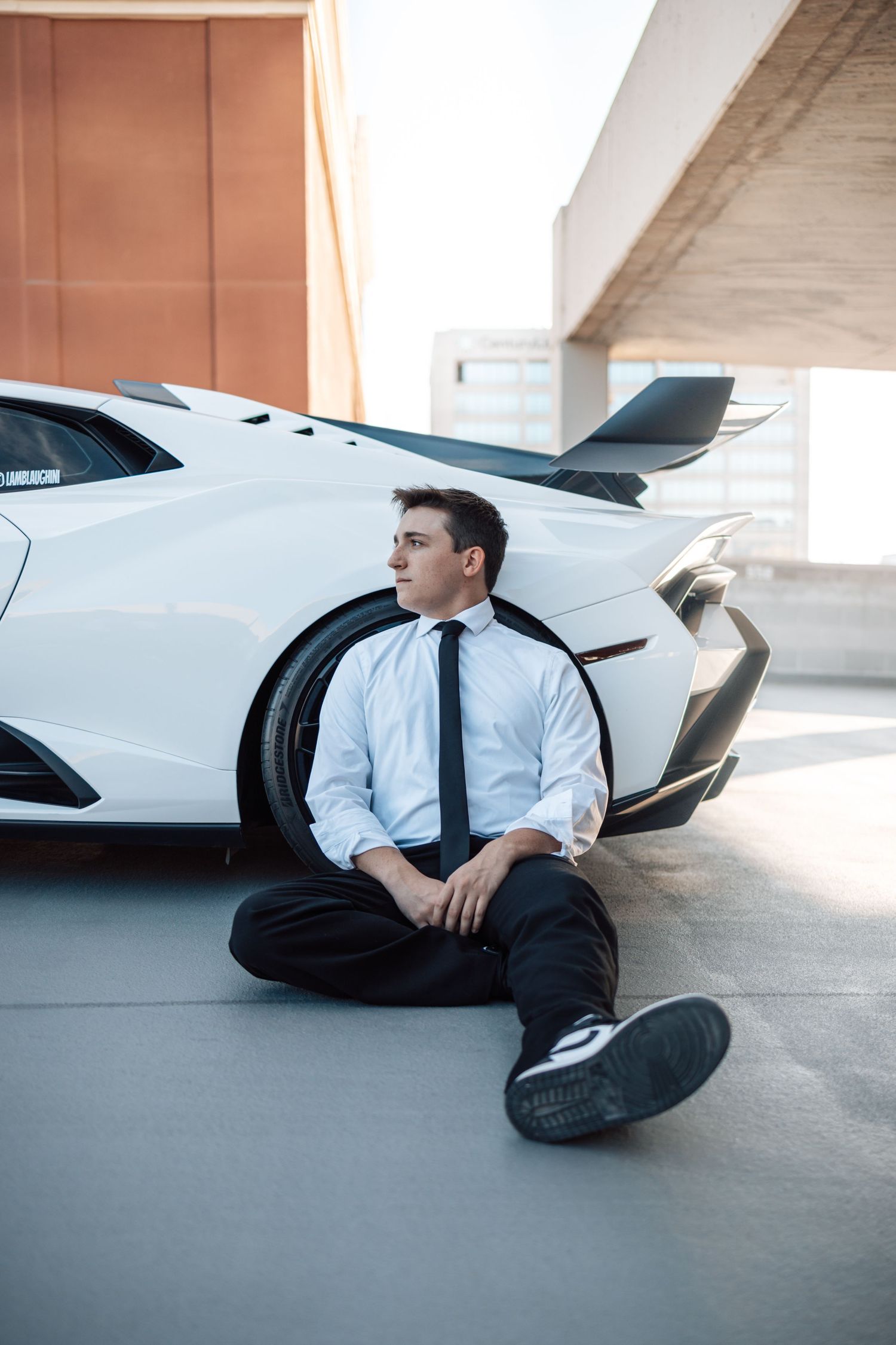 Professional in business attire sits beside a luxury white sports car in a minimalist parking structure.