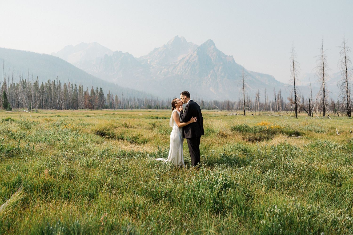 Bride and groom share a romantic kiss in a lush green meadow with majestic mountain peaks in the background.