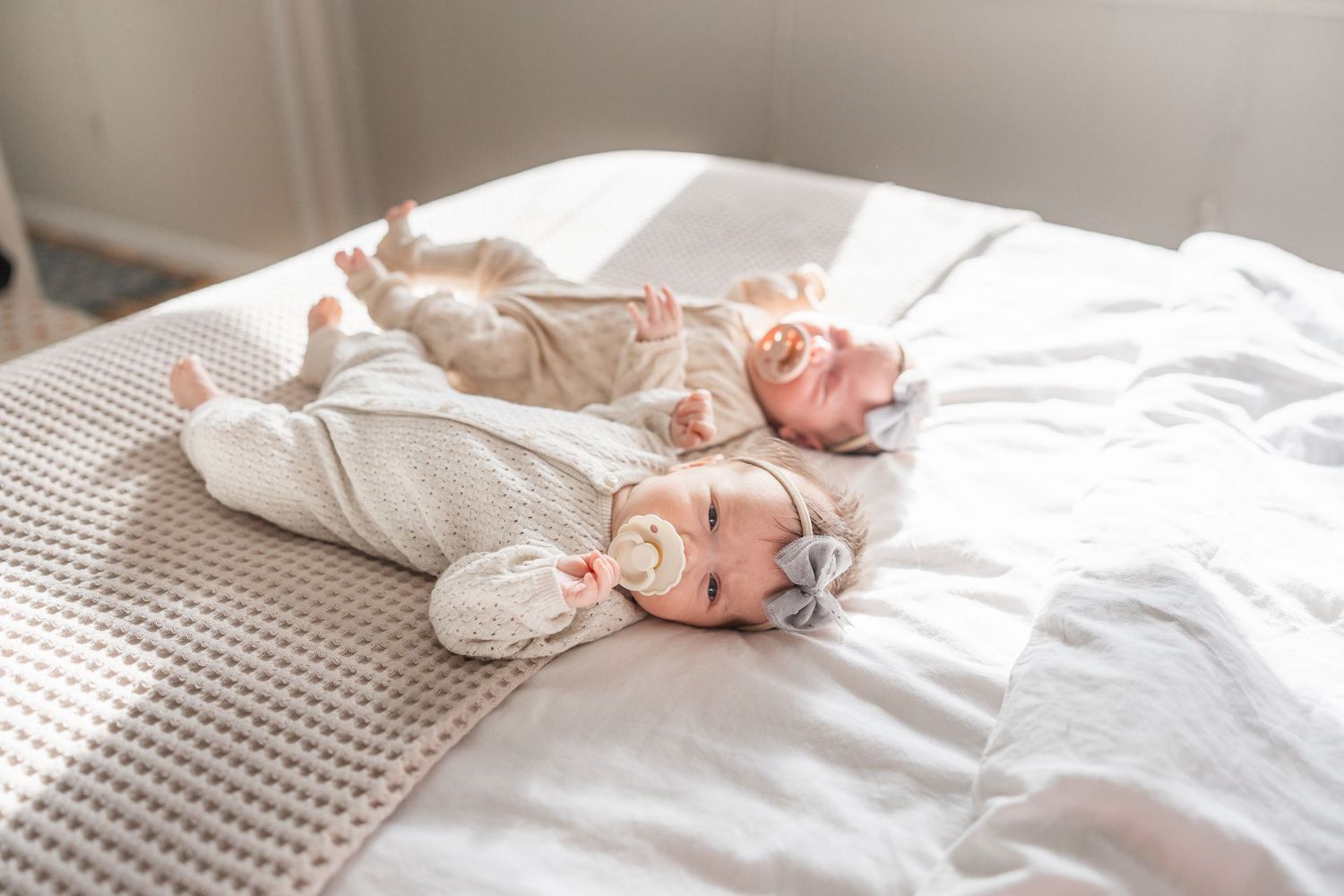 A baby lies on white bedding while wearing a gray onesie and smiling playfully.