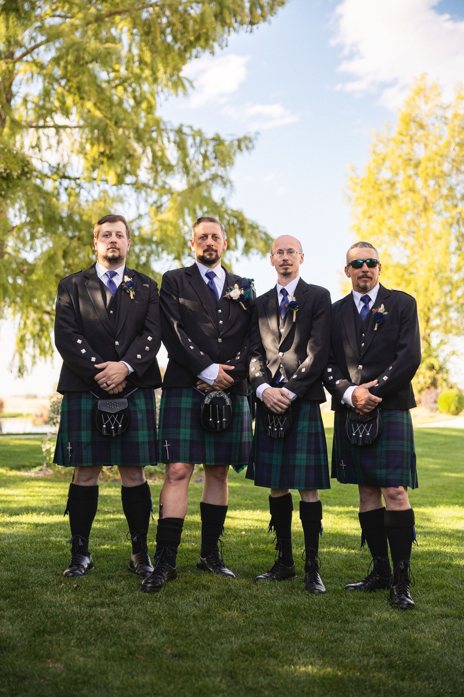 Four groomsmen in matching black jackets and kilts stand in a row outdoors.