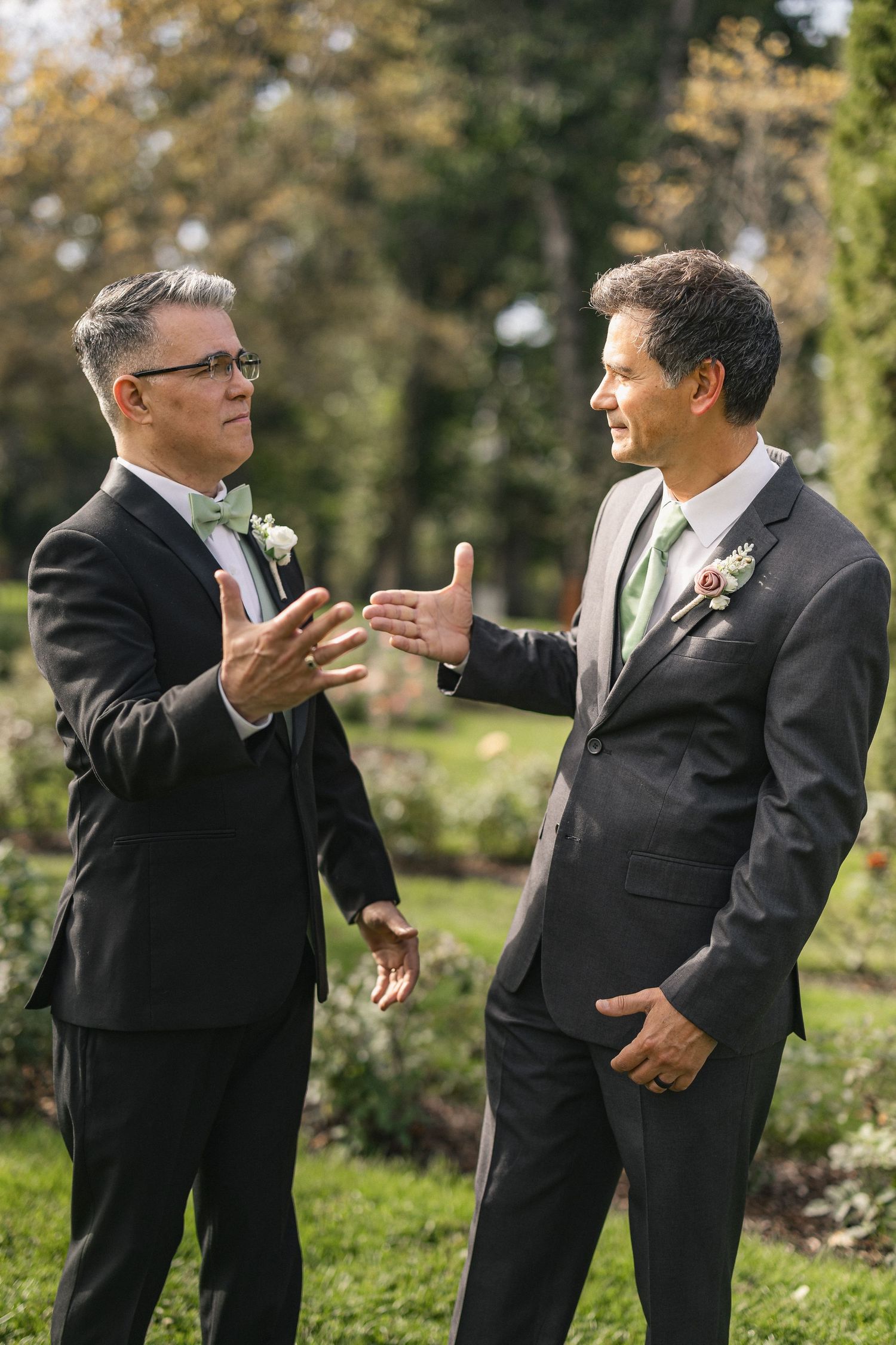 Two men in formal suits engage in animated conversation during outdoor wedding festivities.