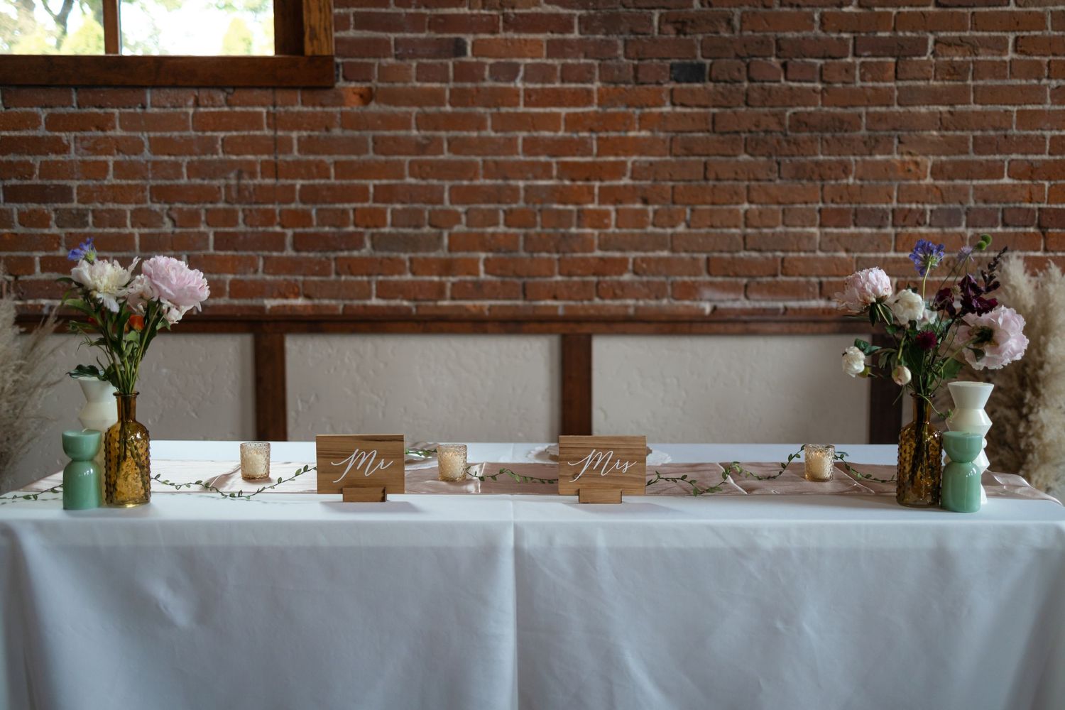 Reception table decorated with white linens, floral centerpieces, and decor against exposed brick wall.