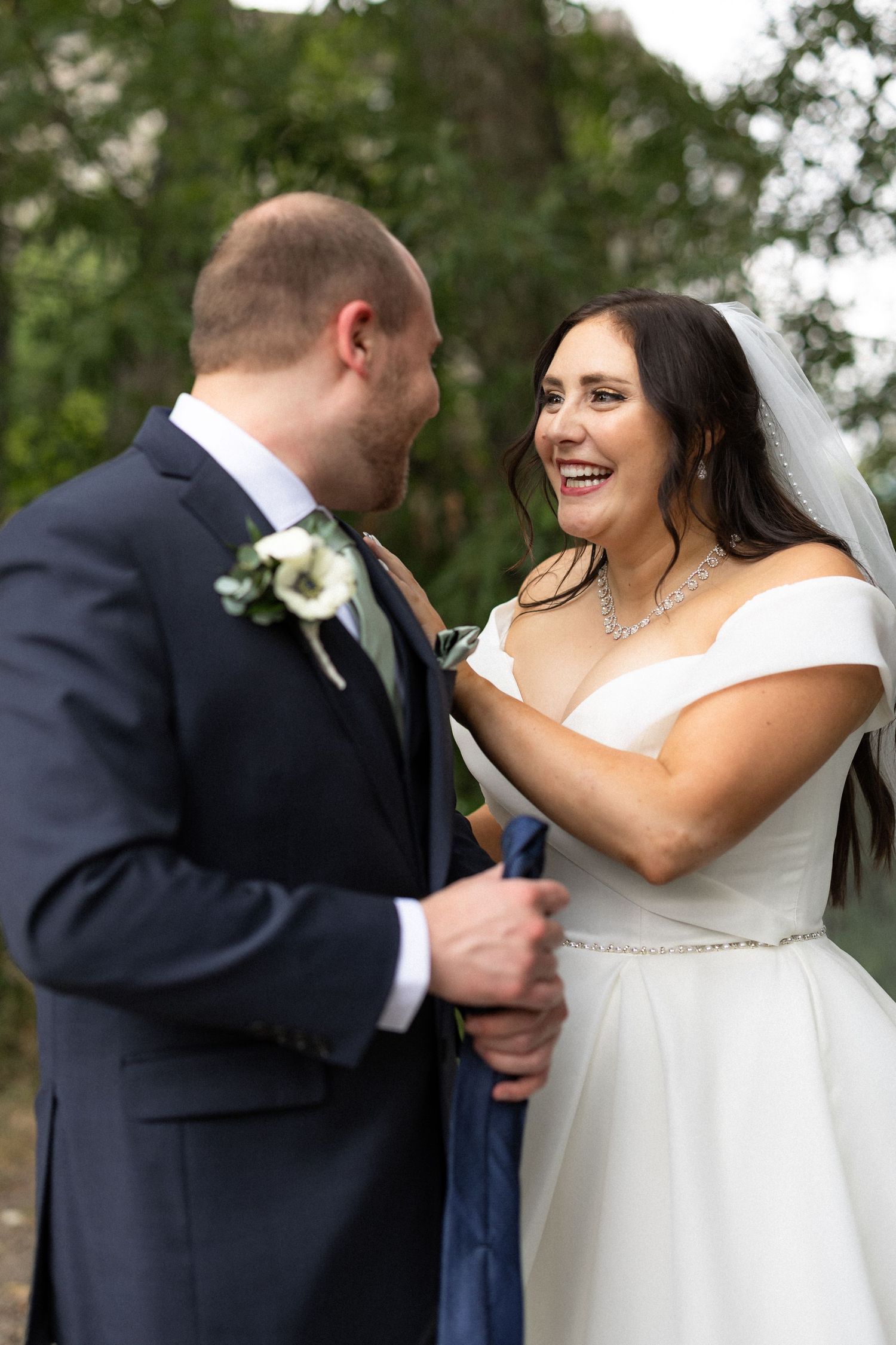A joyful wedding couple shares a playful moment during their first look in a wooded setting.