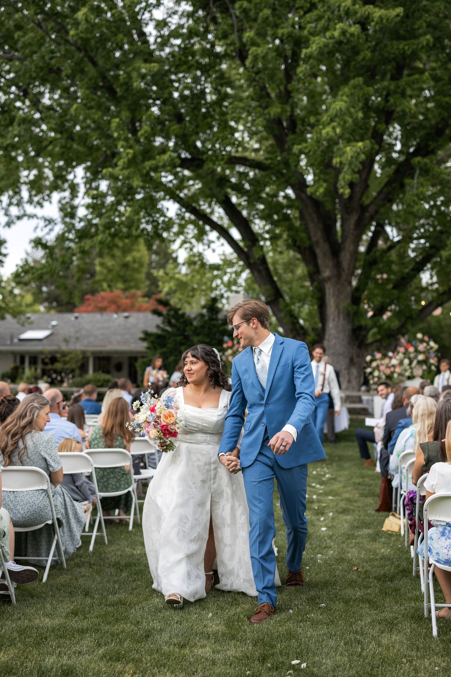 Newlyweds walk down aisle at outdoor garden reception with guests seated in white chairs.