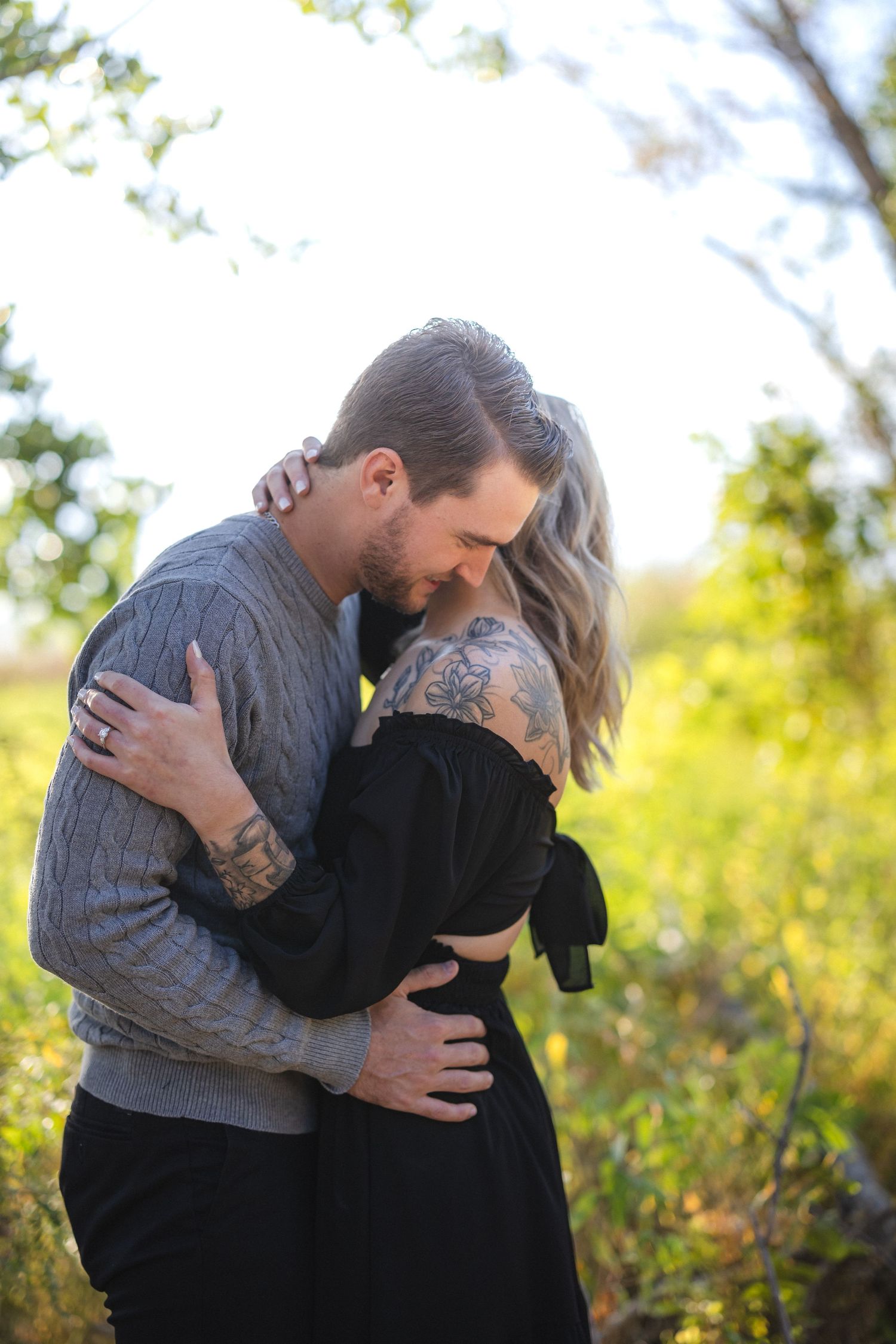 Couple in gray sweater and black dress embrace during sunset photoshoot.
