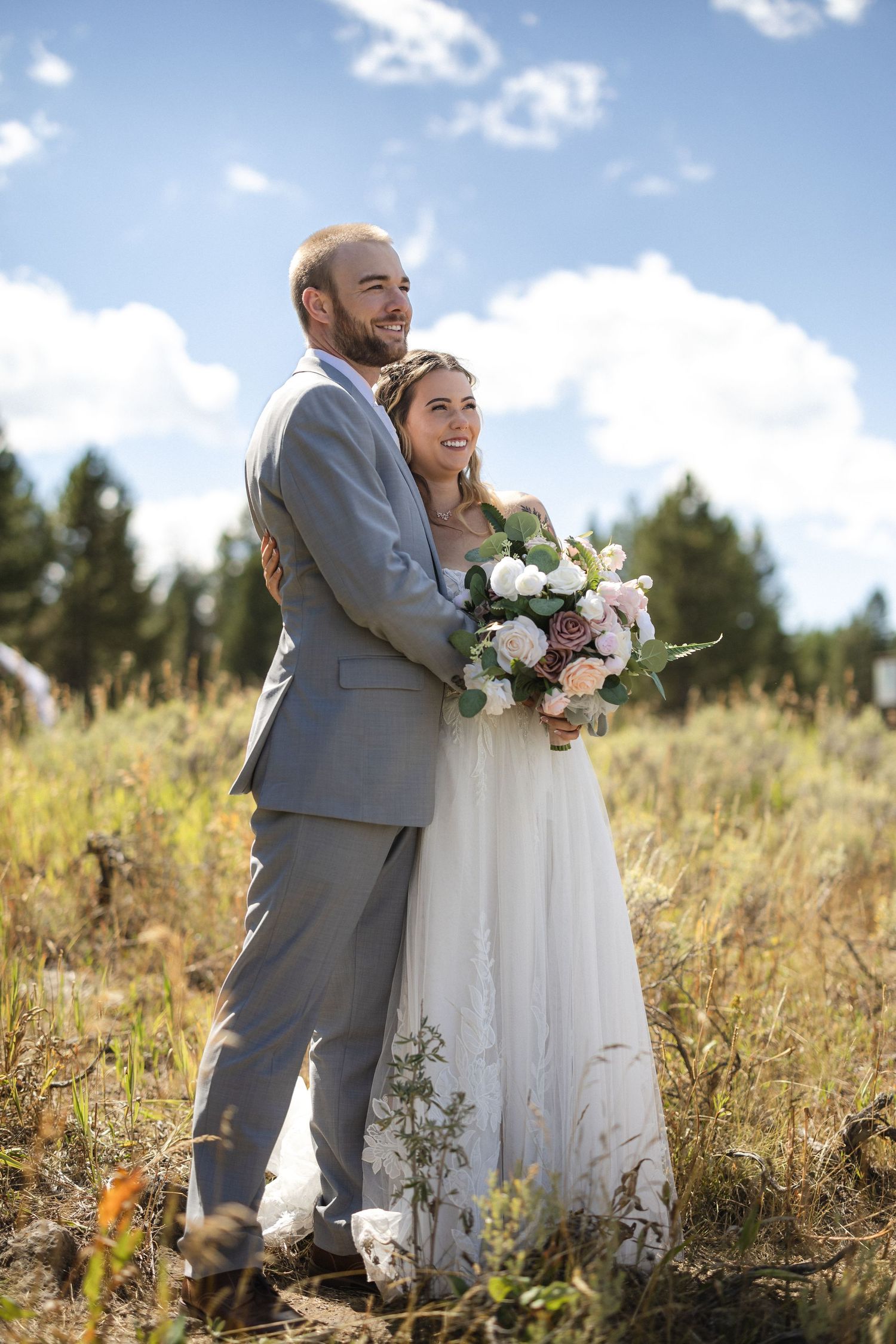 Couple in wedding attire embraces in a sun-lit meadow with white bouquet.