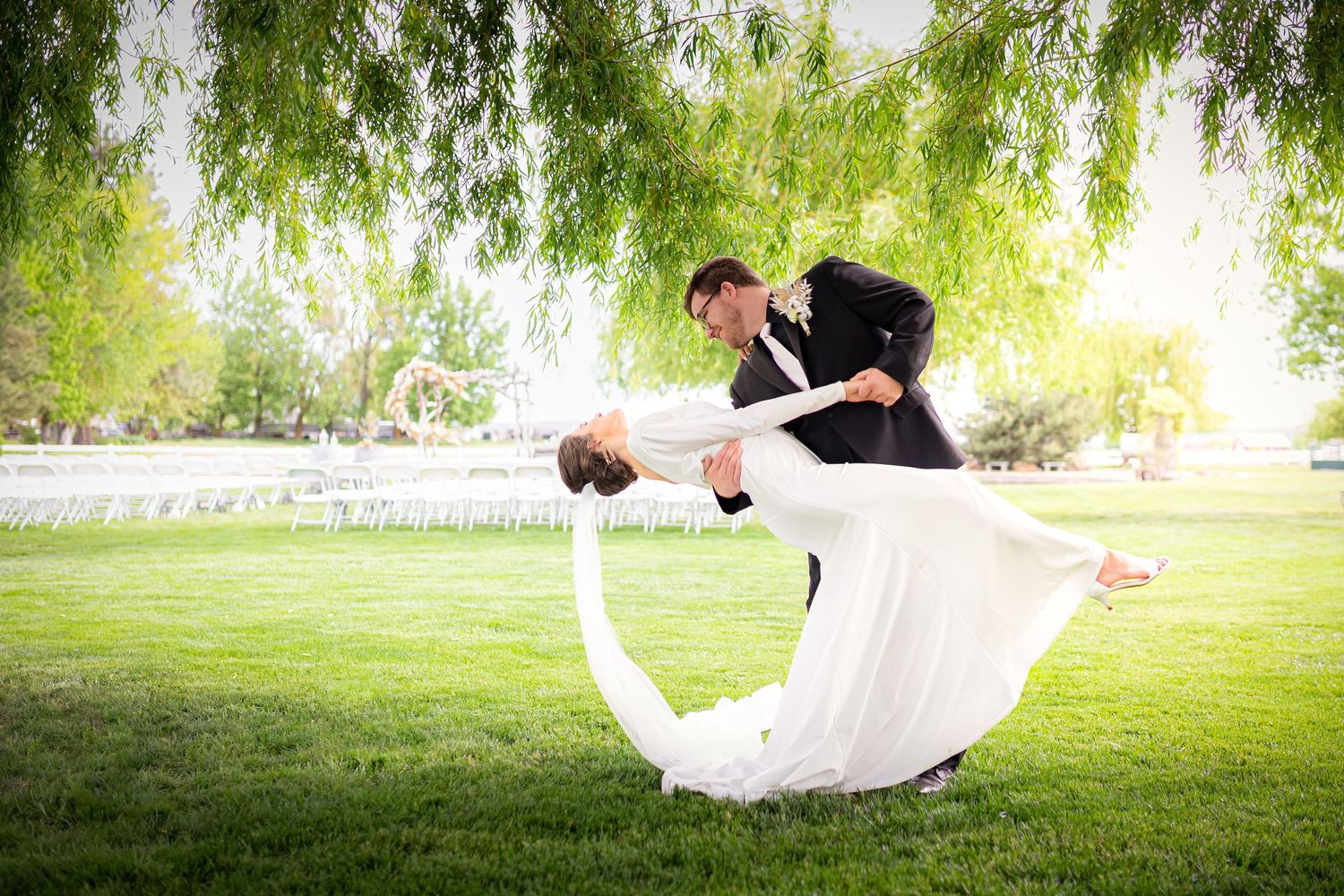 Romantic dip pose of couple on green lawn under weeping willow branches.