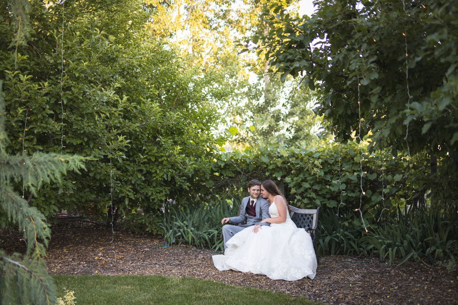 Newlyweds share an intimate moment on a garden bench surrounded by greenery.