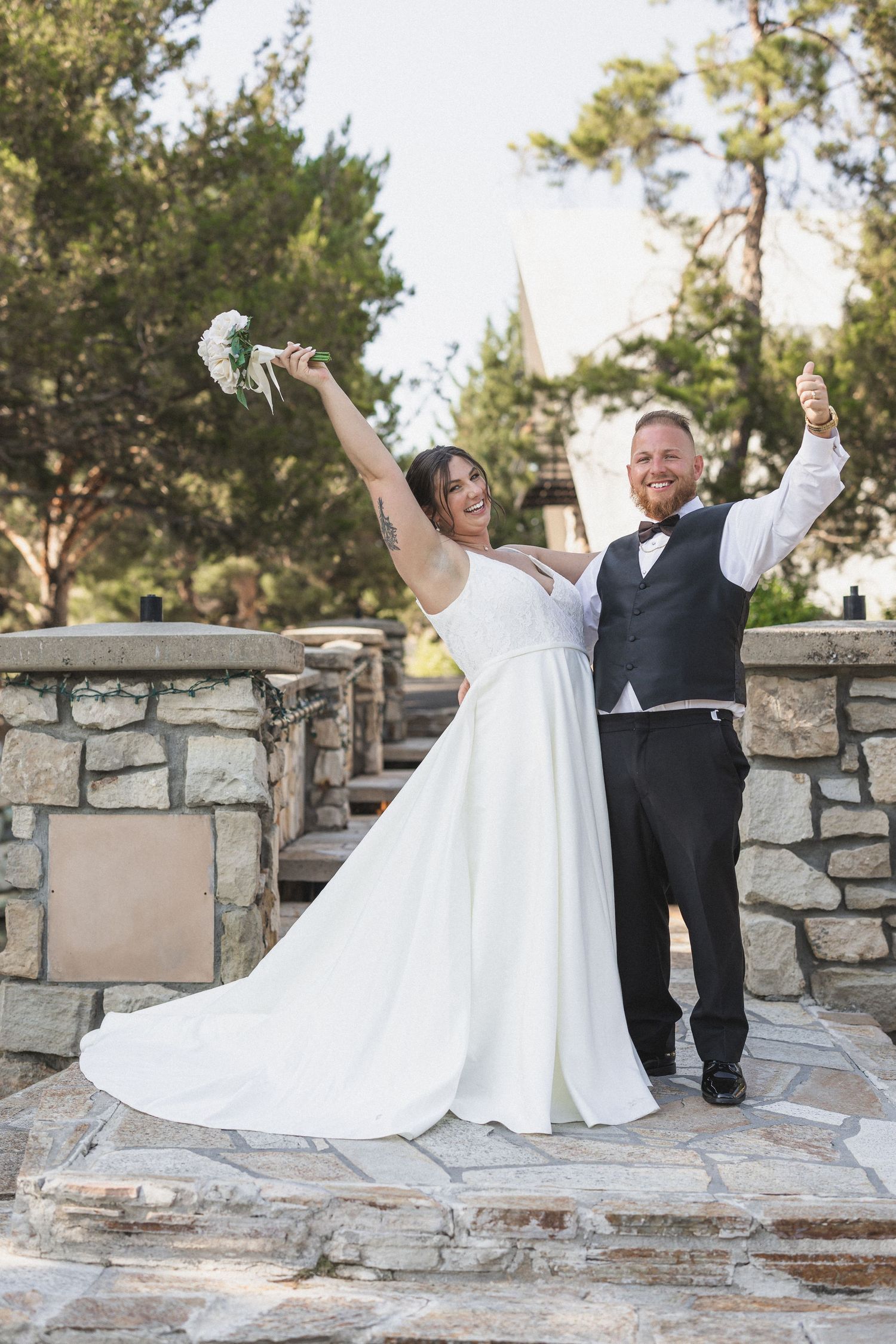 Joyful newlyweds celebrate with raised arms on a stone patio, bride in flowing white dress holding bouquet.